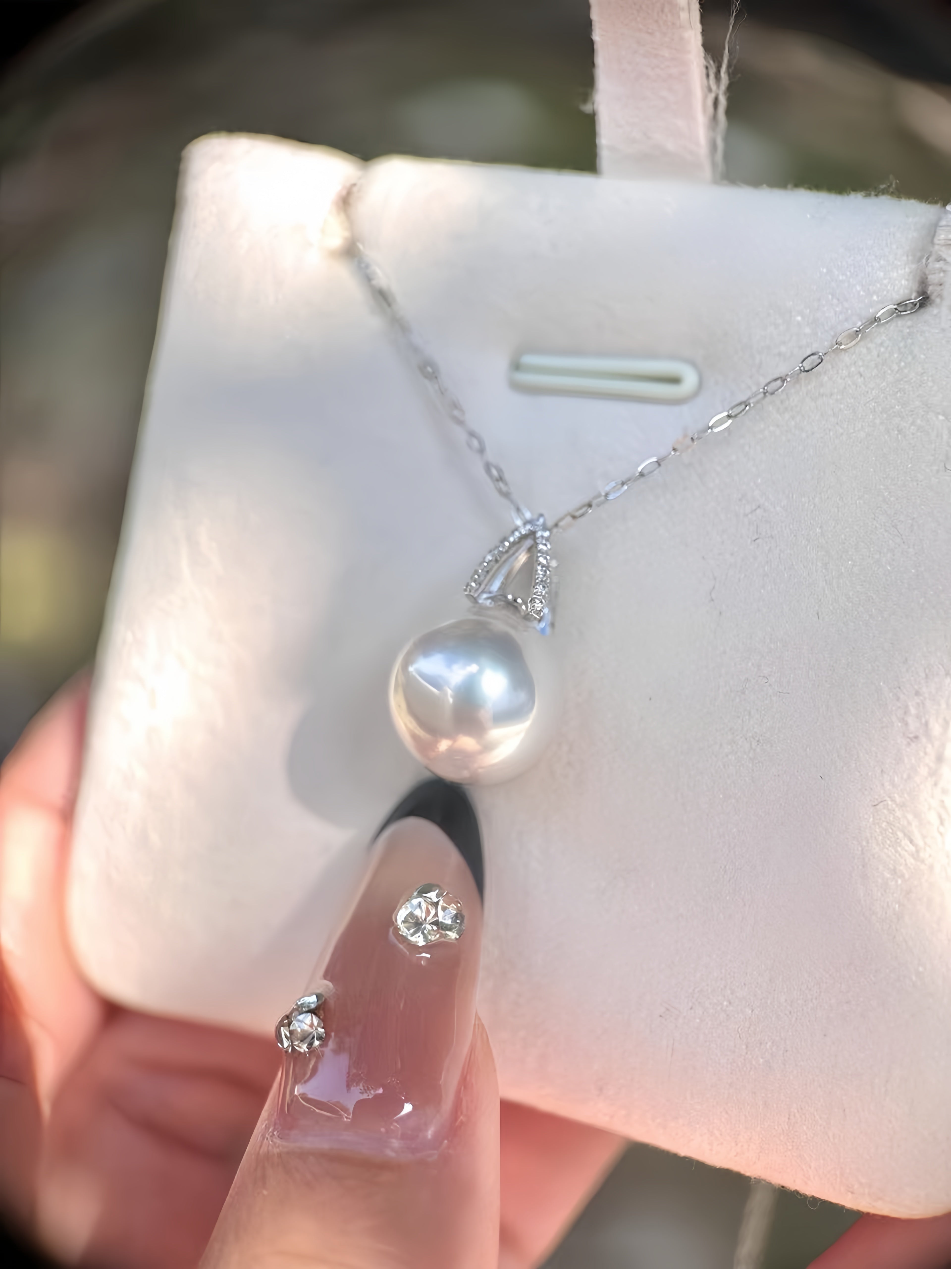 A silver necklace with a large white freshwater pearl pendant displayed on a white jewelry card, held by a hand with decorated nails.