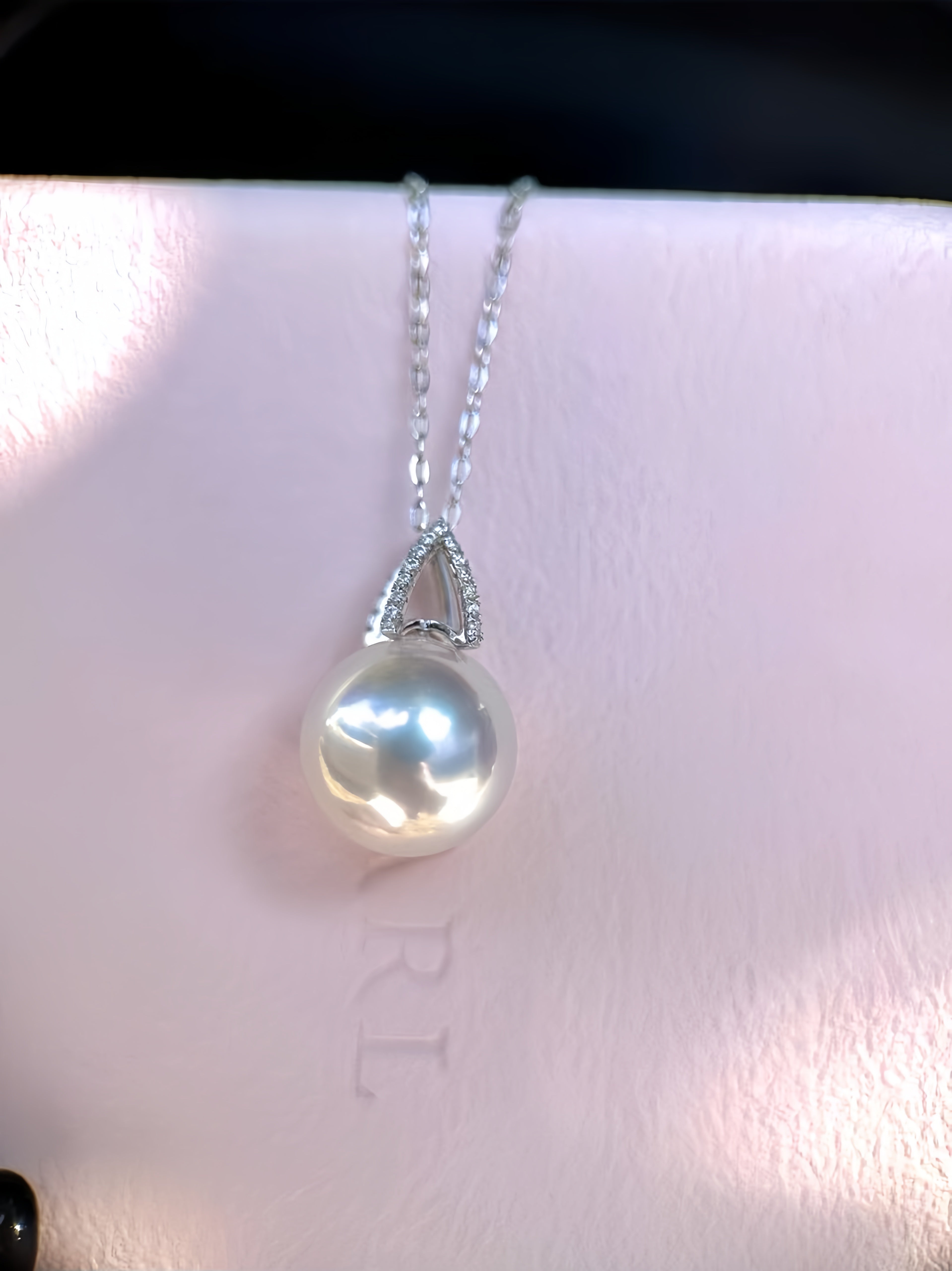 A silver pendant necklace featuring a single lustrous white freshwater pearl and a small halo of clear stones, resting on a light pink textured surface.