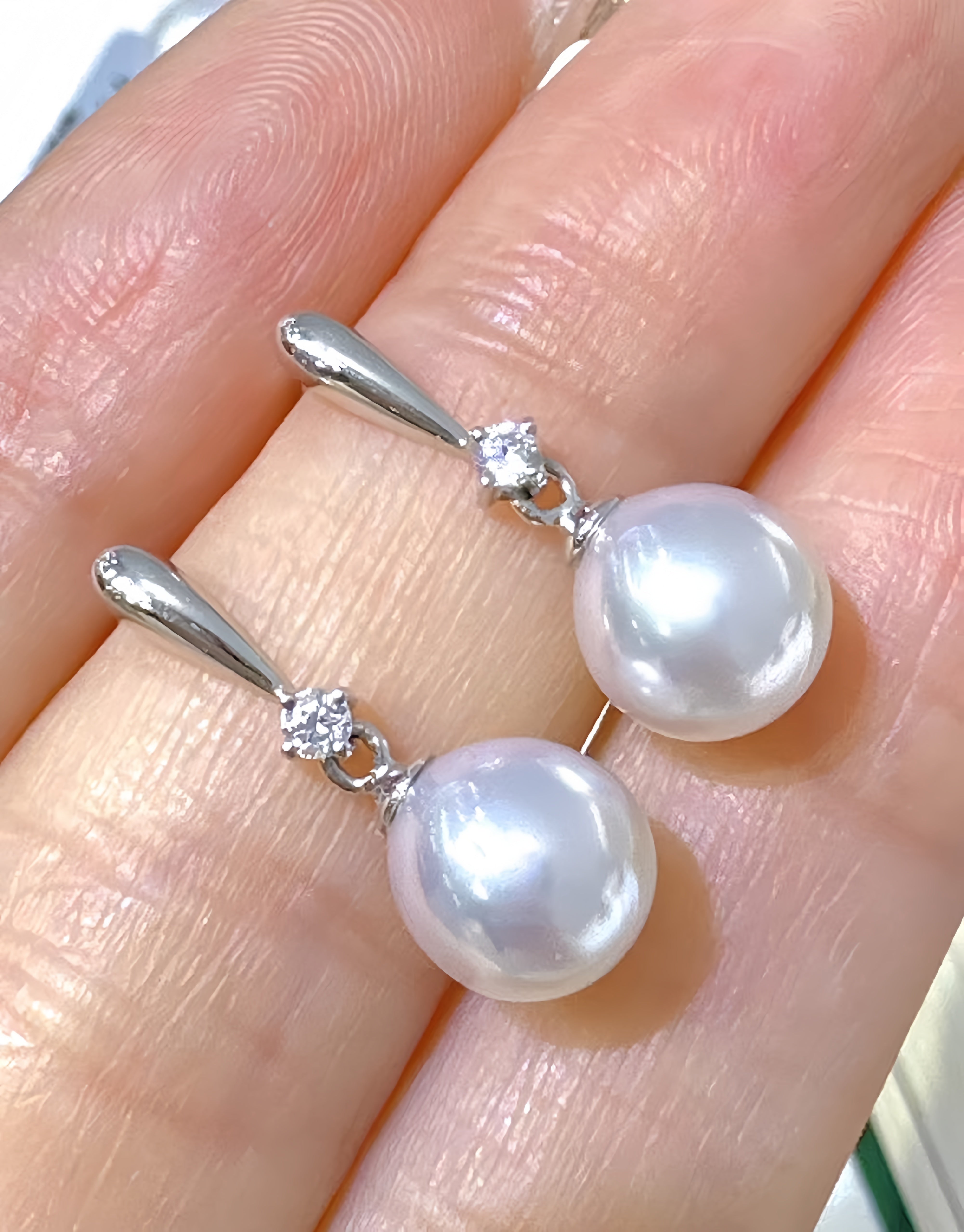 A pair of silver drop earrings with large white pearl-like round drops and small clear stones, shown resting on a person's fingers.
