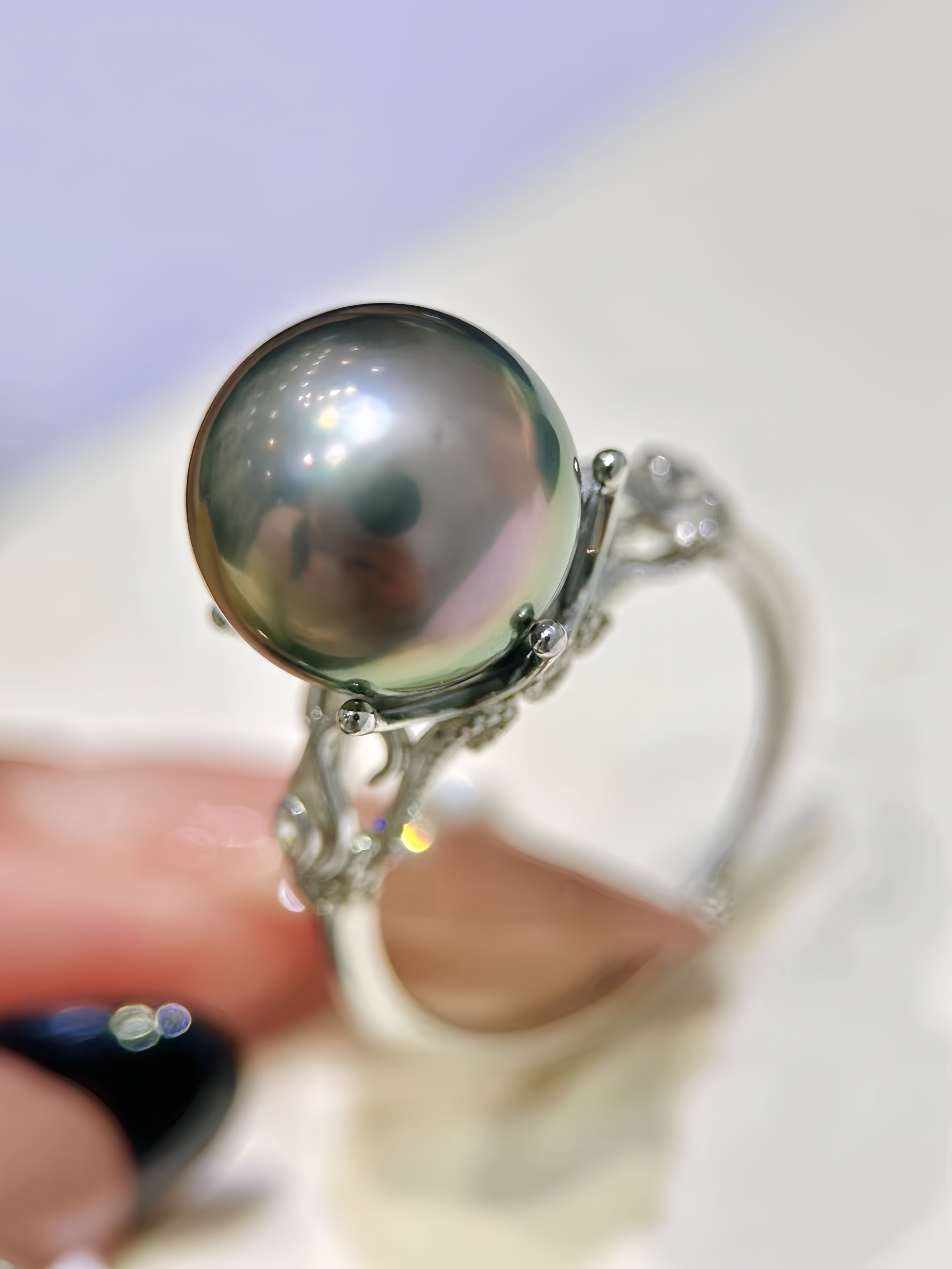 A close-up of a silver ring featuring a large, round, dark gray pearl with iridescent hints, held by fingers against a soft-focus light background.