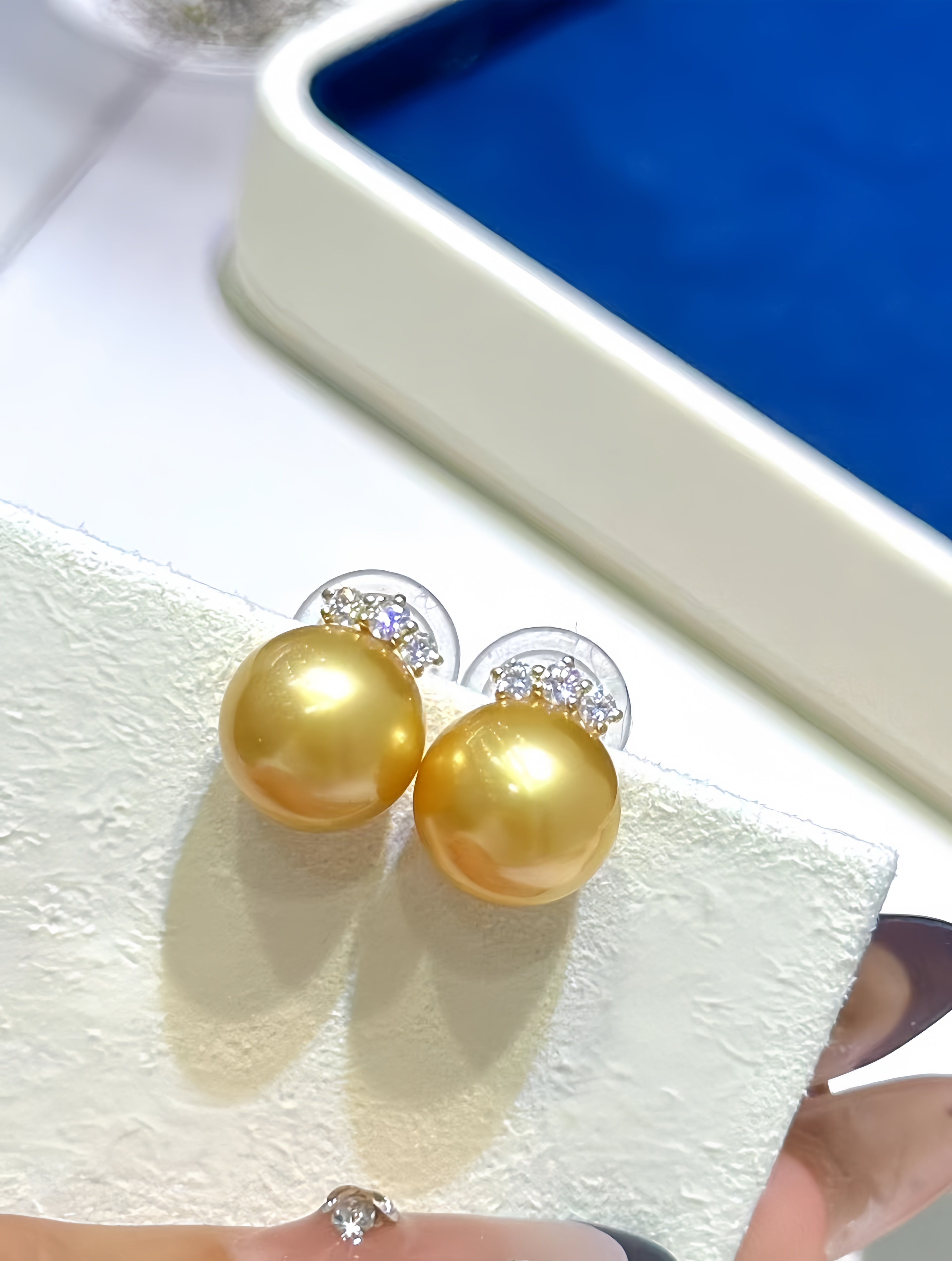 A pair of gold pearl stud earrings with small clear crystal accents on a textured white display block, with a hand holding the corner and a blue jewelry box in the background.