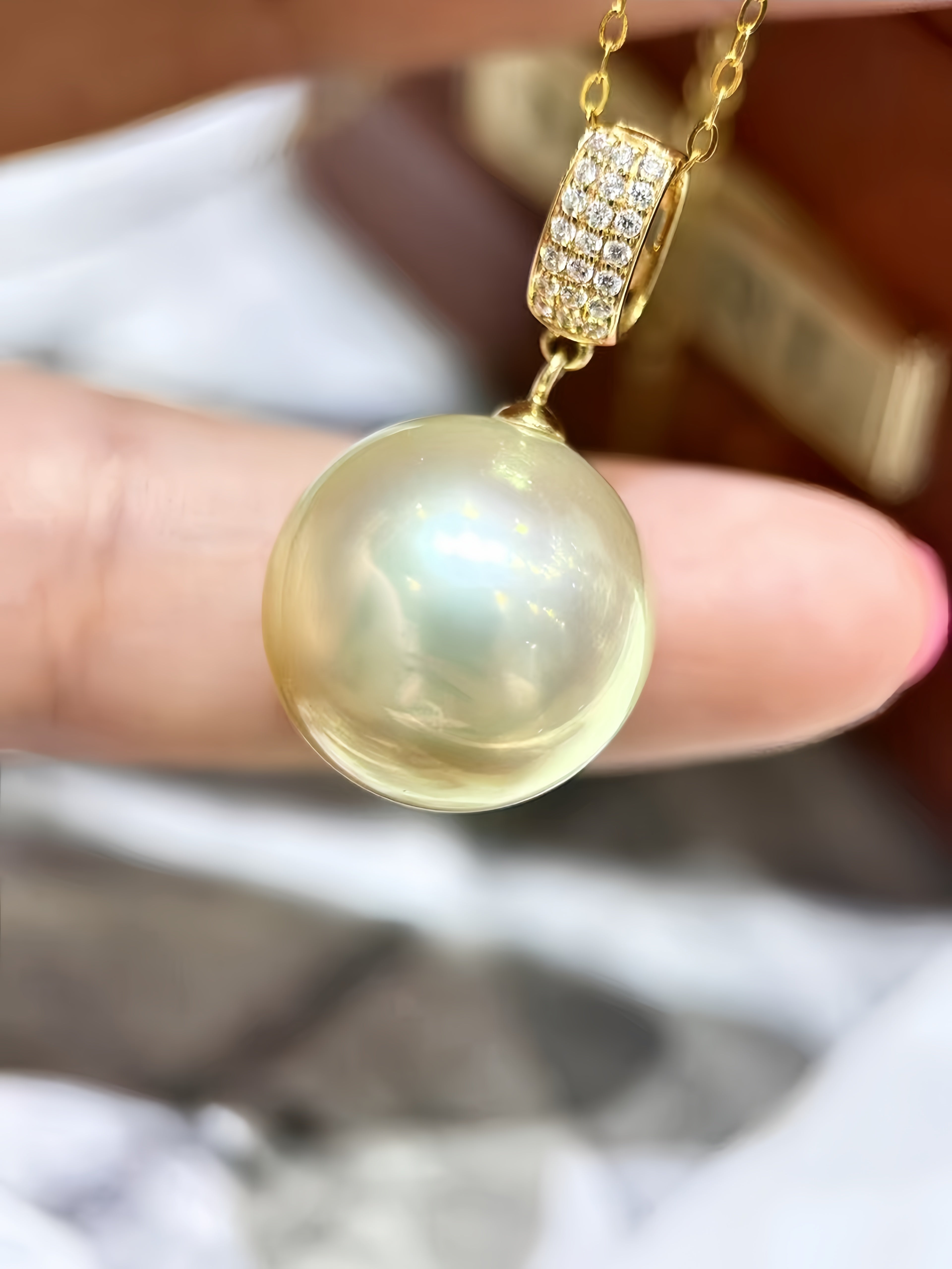 A close-up of a gold pendant necklace featuring a large, round, lustrous pearl-like bead and a gold clasp adorned with small clear stones, held between fingers.