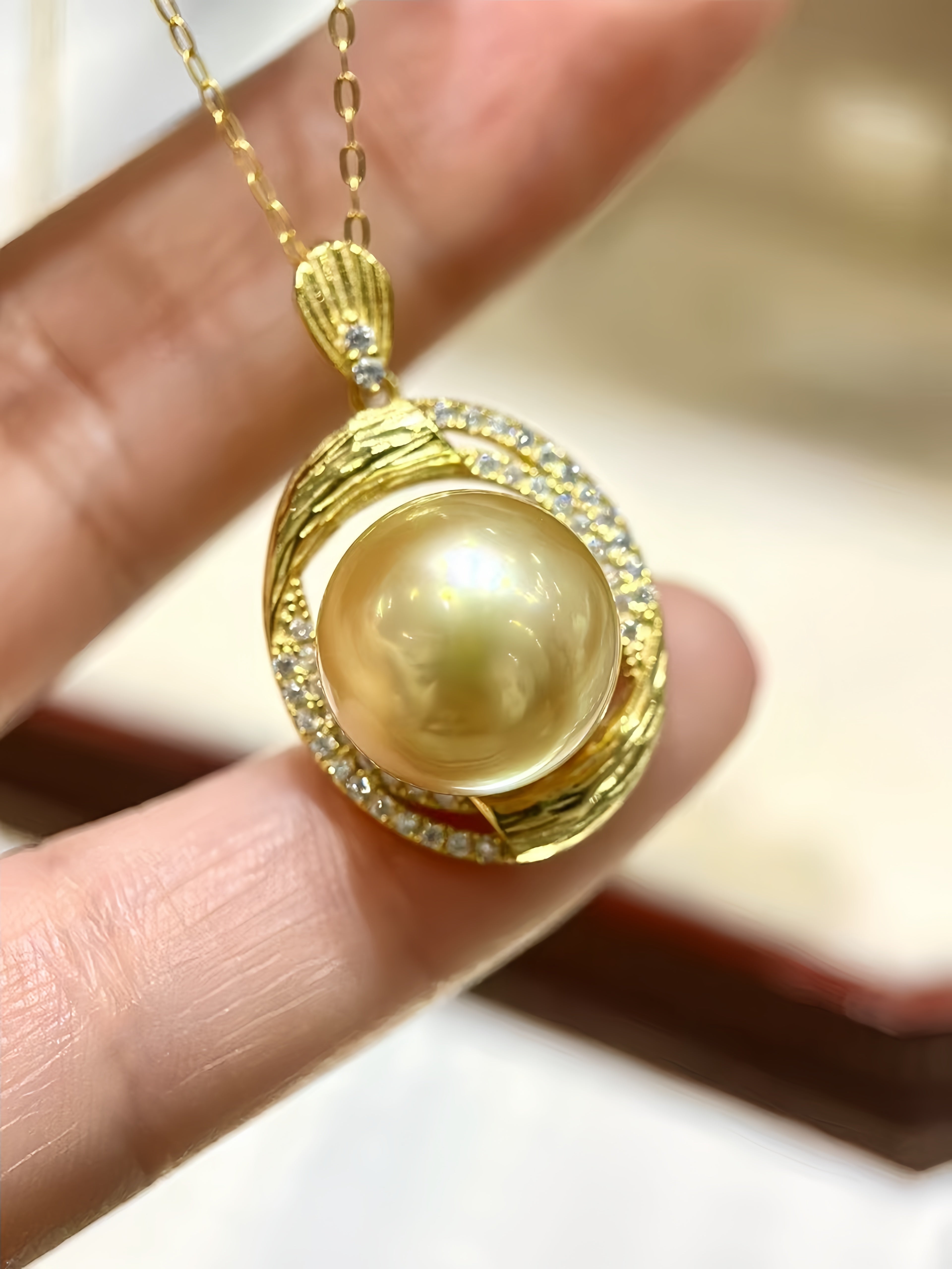 A gold pendant necklace with a large round yellow pearl surrounded by small clear stones, held in a person's fingers against a softly blurred light background.