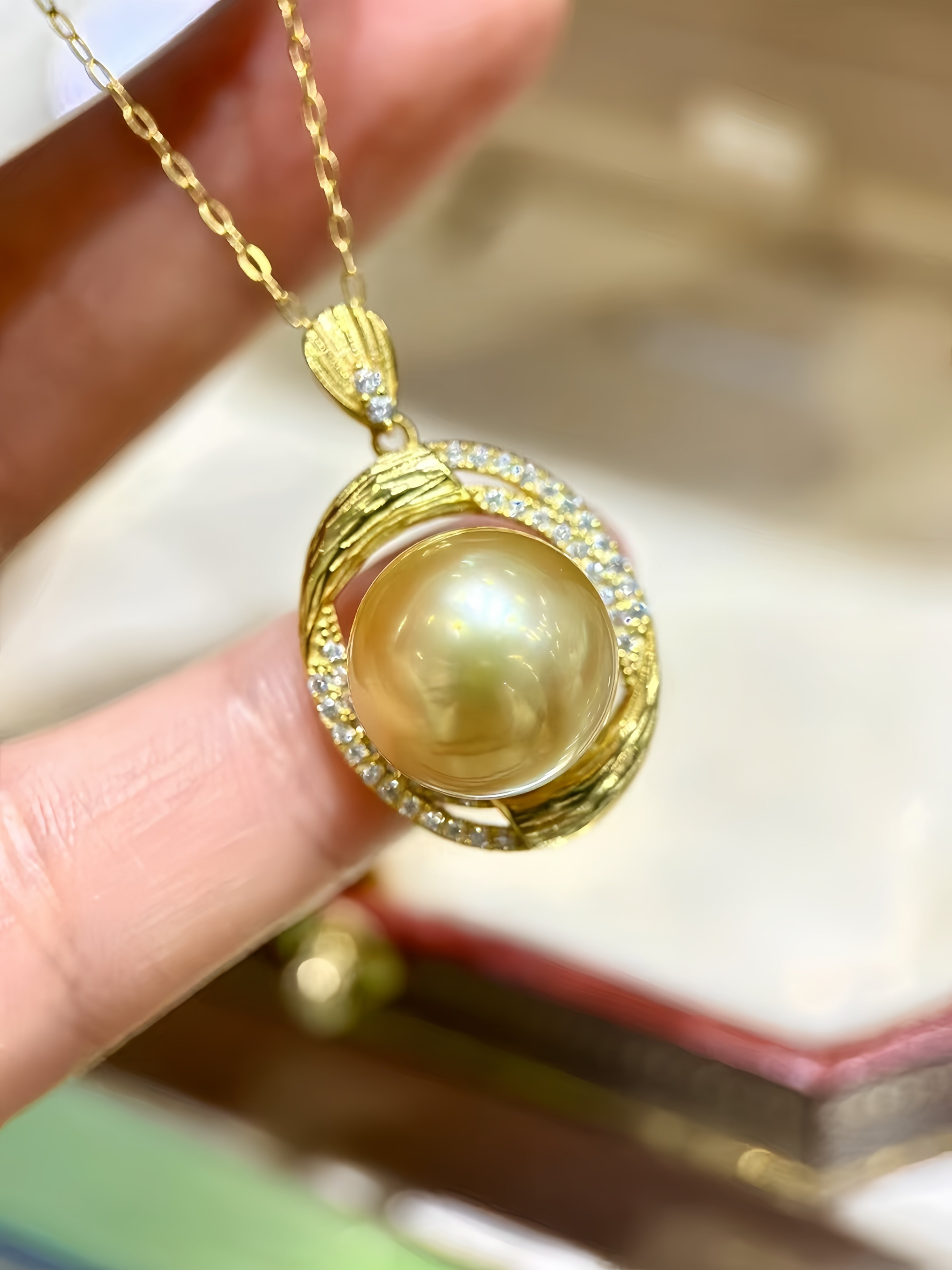A gold pendant necklace featuring a large round pearl surrounded by small clear stones and textured gold details, held by a hand against a softly blurred background.