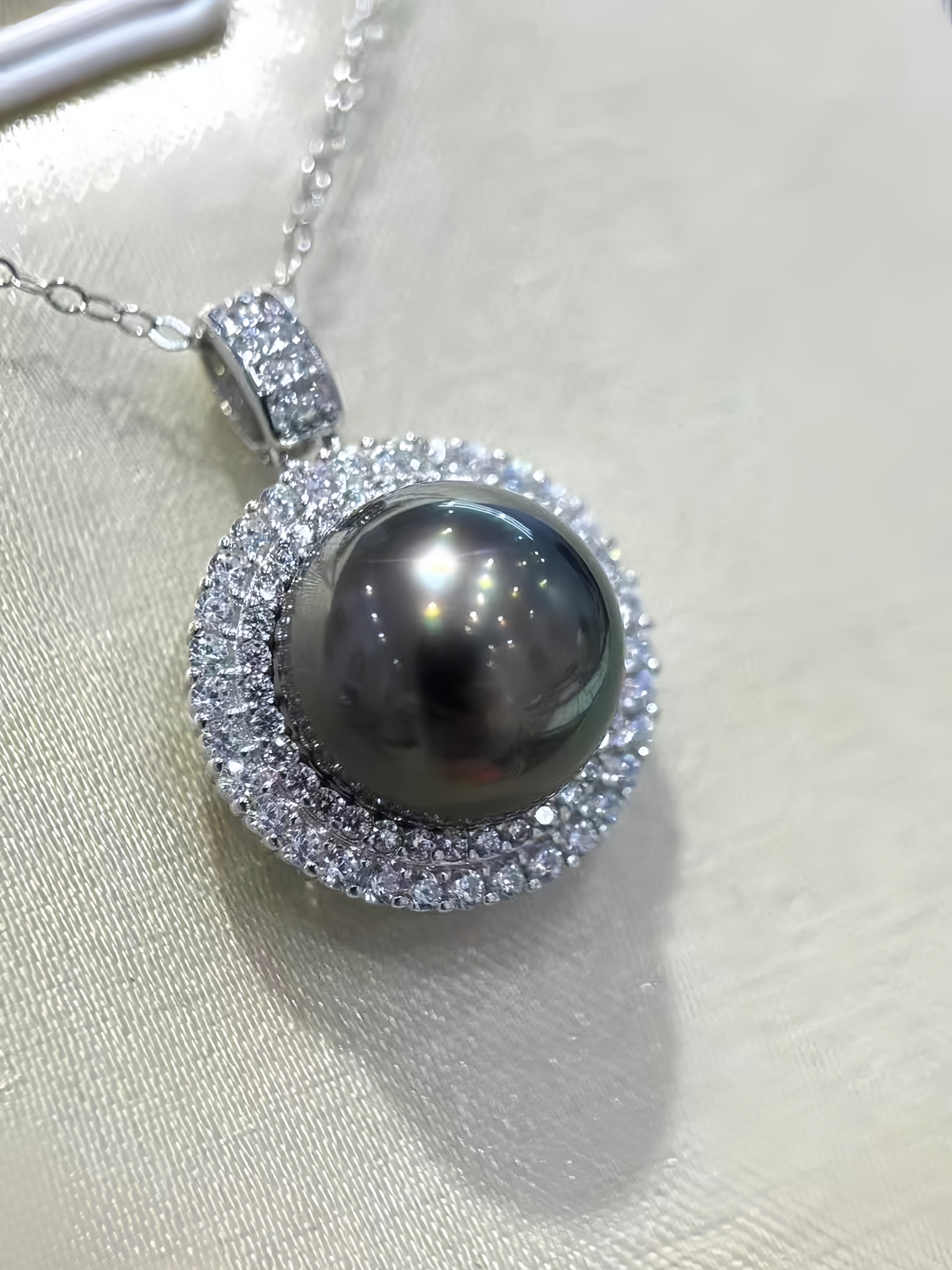 A close-up of a silver pendant necklace featuring a large, dark gray pearl surrounded by a halo of small clear crystals, resting on a light beige textured fabric surface.