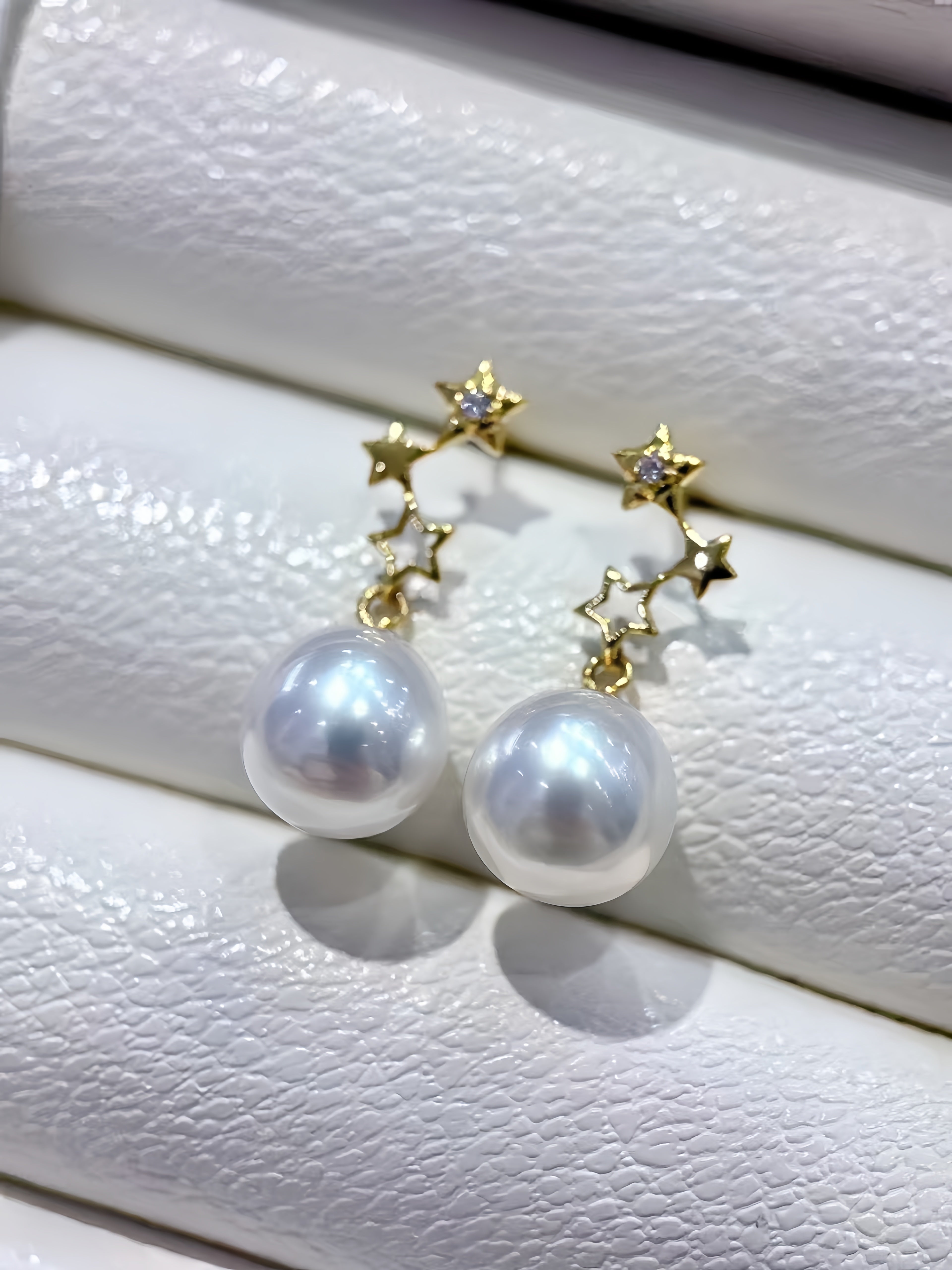 A pair of gold star-shaped dangle earrings with large white freshwater pearls, displayed on a textured white jewelry stand.