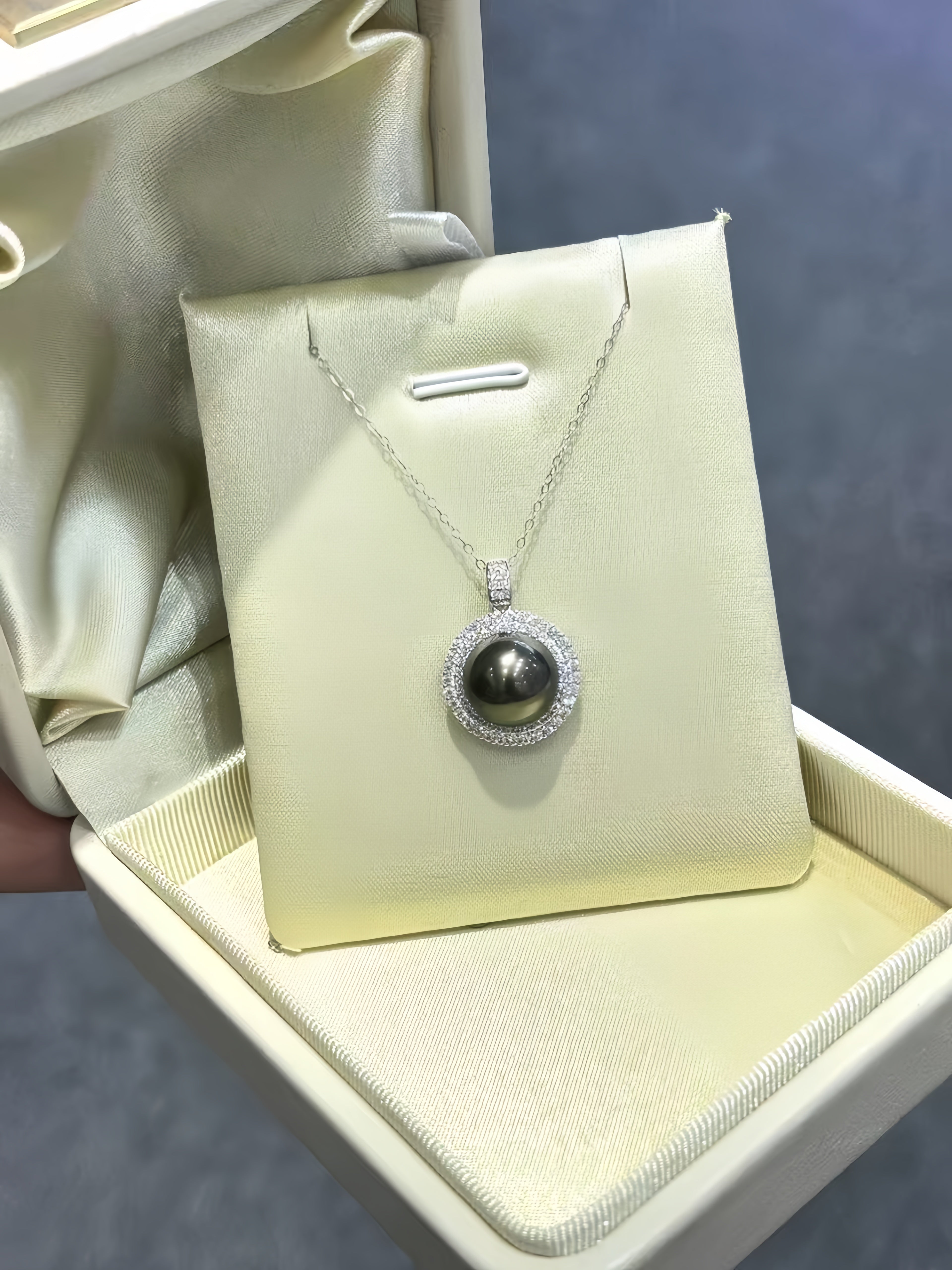 A silver pendant necklace featuring a dark gray pearl surrounded by a halo of small clear stones, displayed on a cream-colored jewelry card inside an open presentation box.