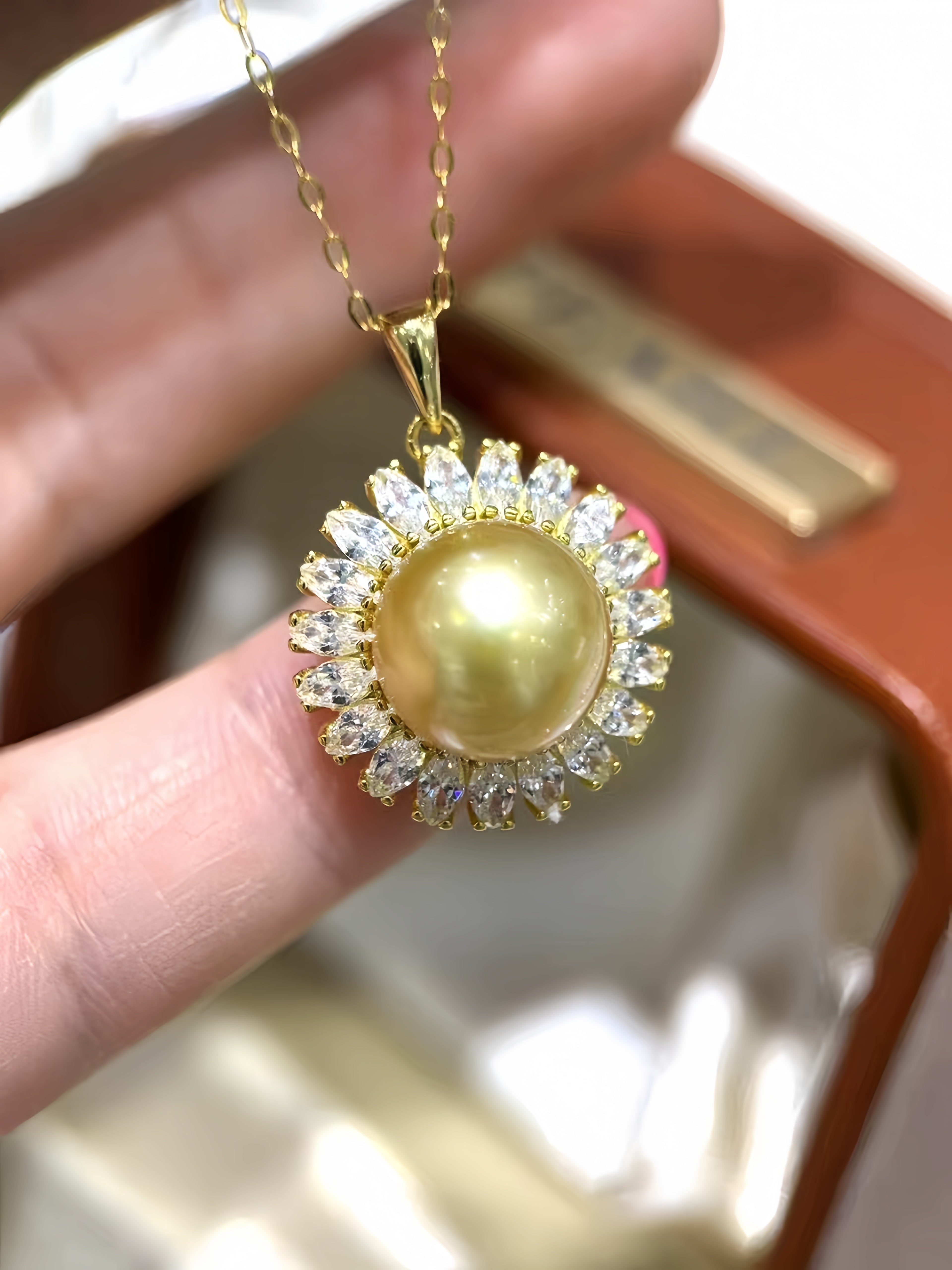 A gold pendant necklace featuring a large round yellow pearl surrounded by a halo of clear crystals, held by a hand in front of a brown jewelry box.