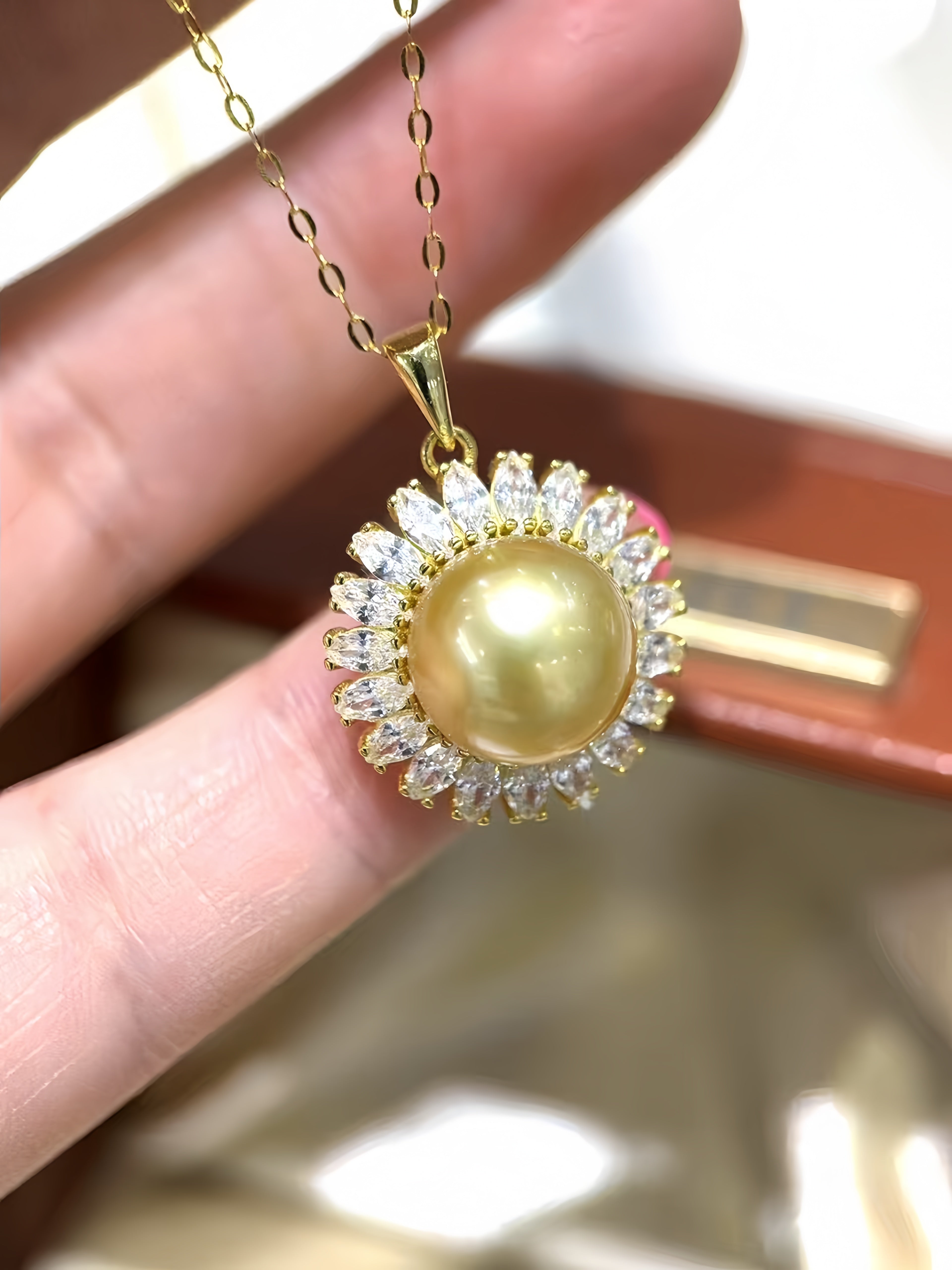 A gold pendant necklace with a large round yellow pearl surrounded by small clear stones, held between fingers.