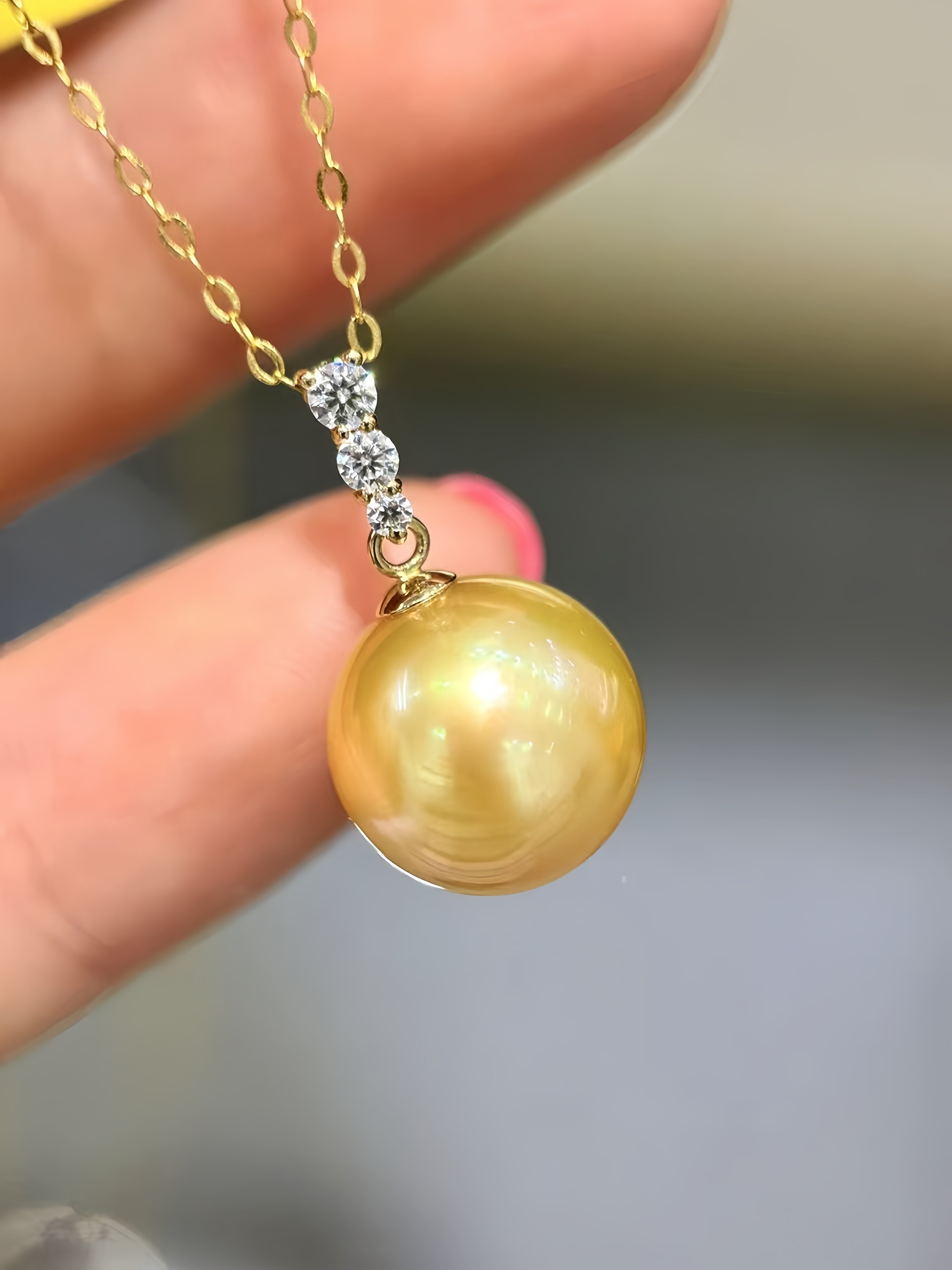 A close-up of a gold necklace with a large round yellow pearl pendant and three small clear stones, held between fingers.