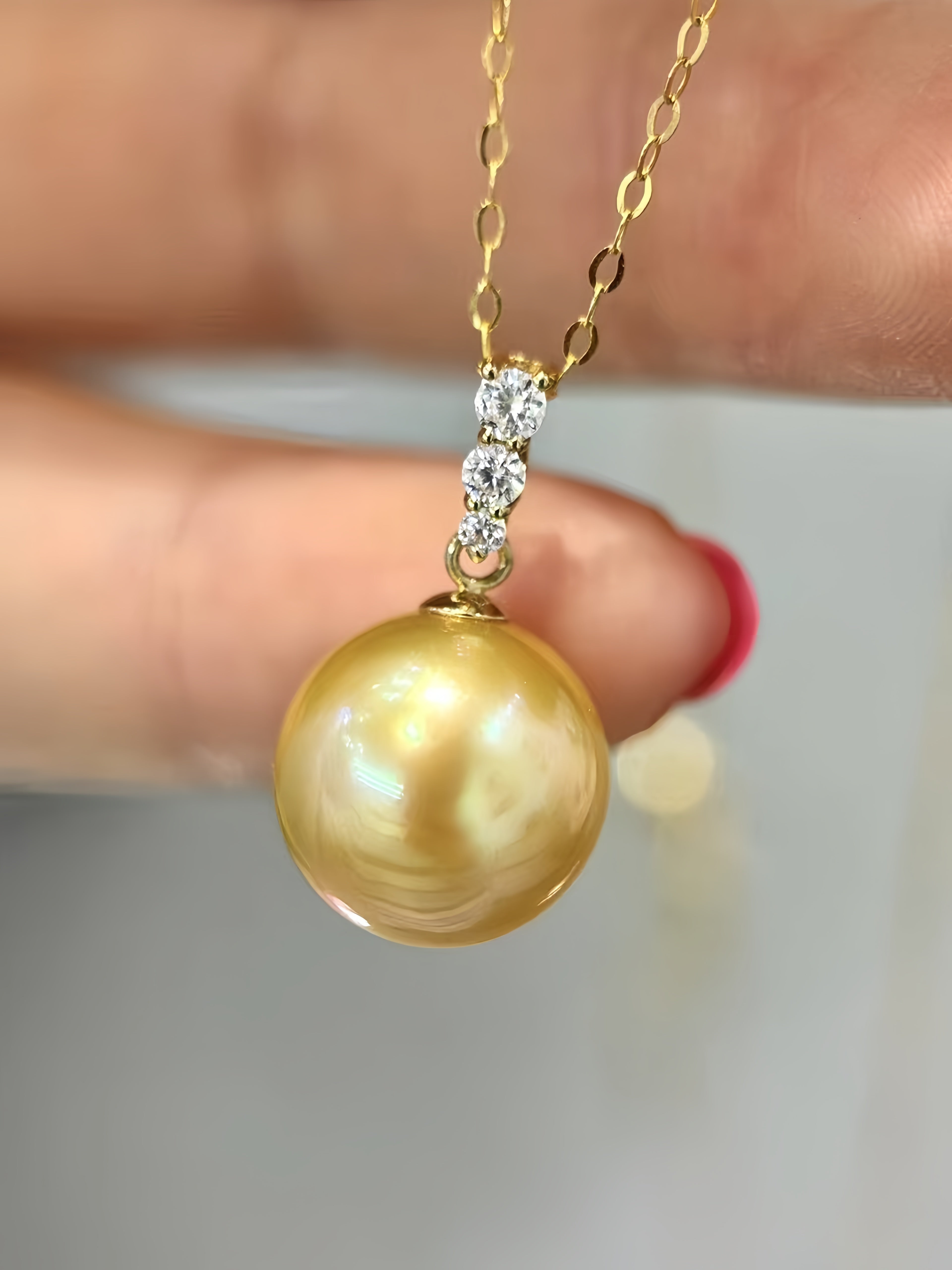 A gold pendant necklace featuring a large, lustrous round pearl pendant and a three-stone diamond setting, held by a hand.
