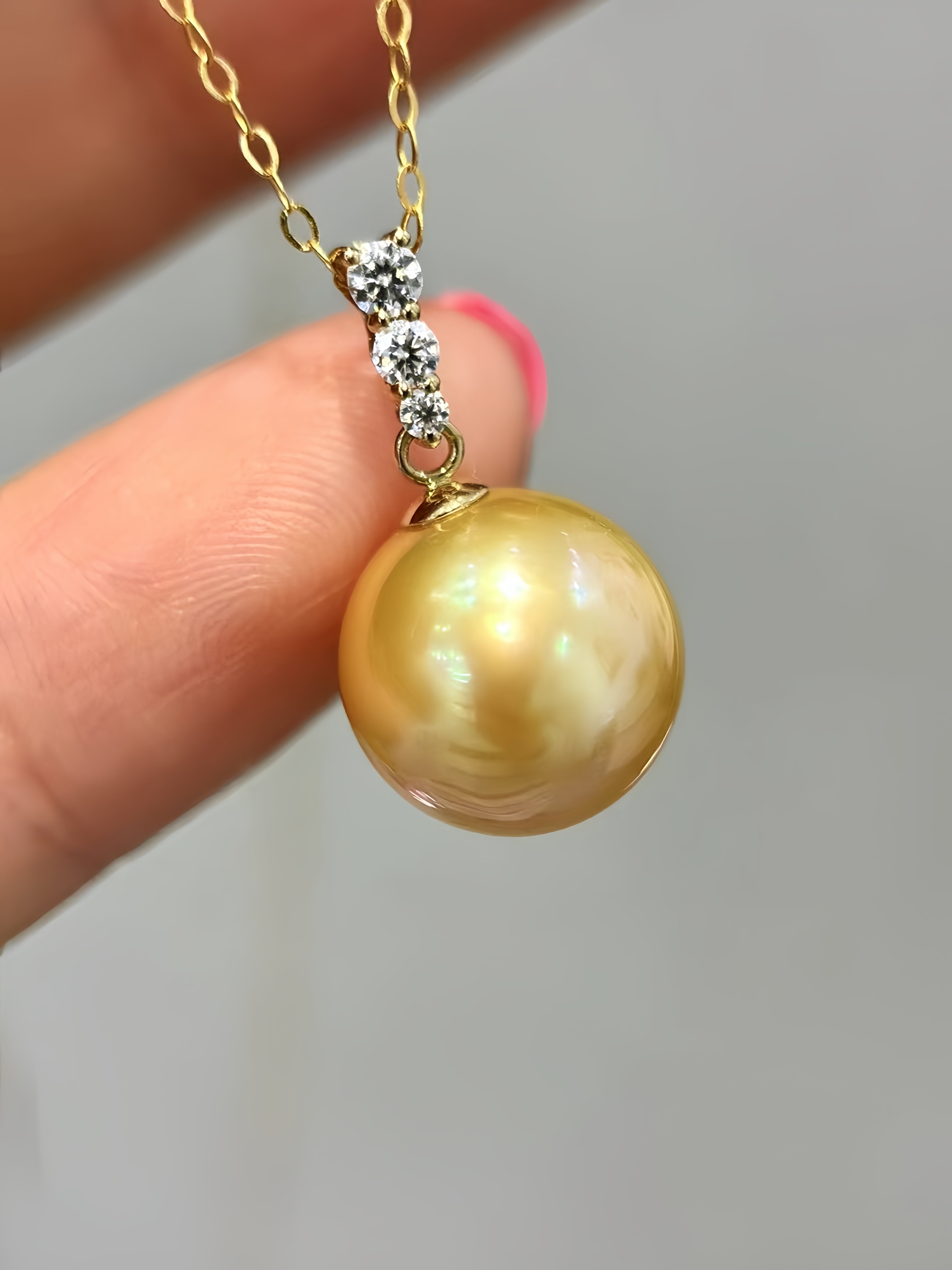 A gold pendant necklace featuring a large, lustrous golden pearl suspended from a gold chain with small clear stones, held by a hand.