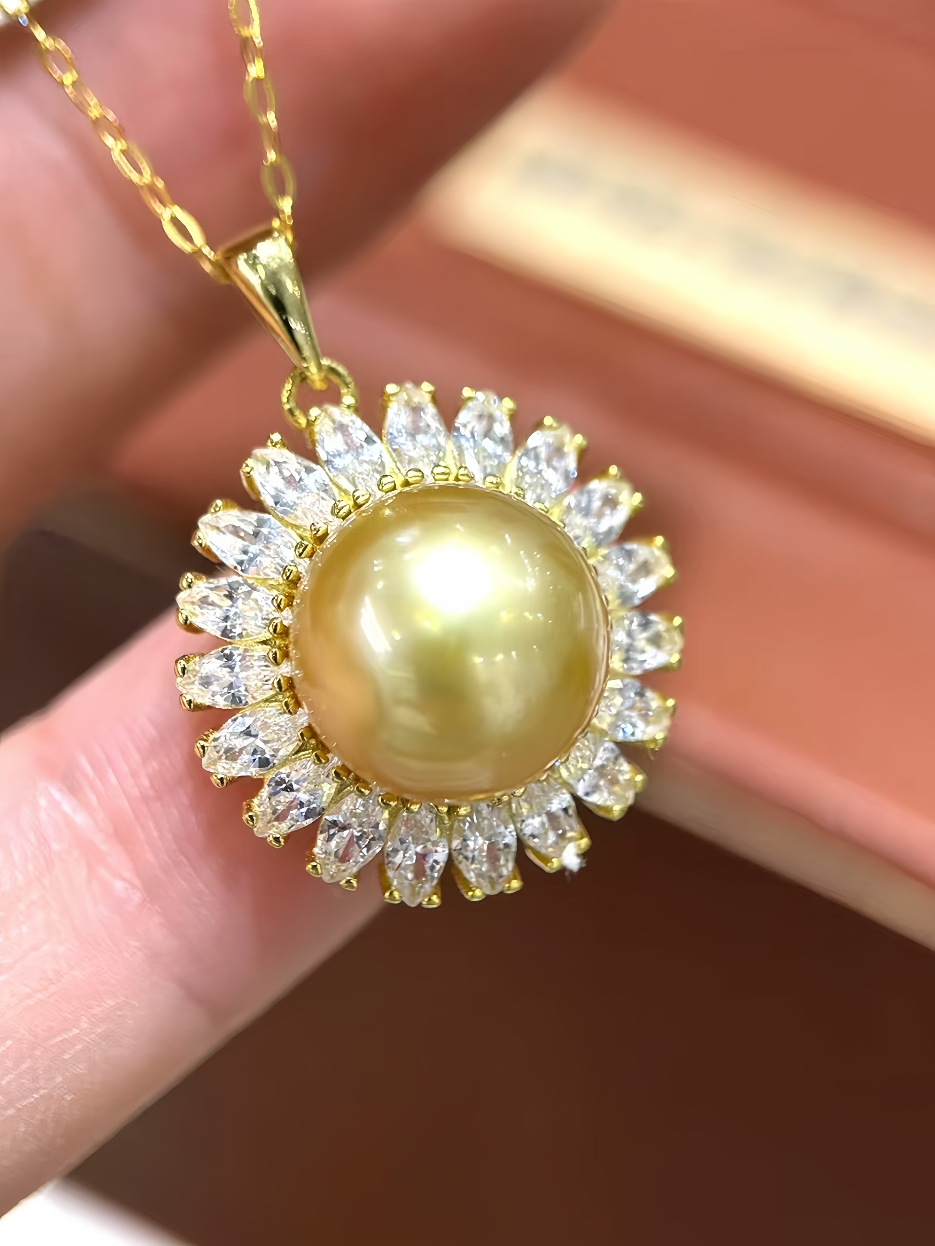 A gold pendant necklace featuring a large round yellow pearl surrounded by a halo of clear baguette-cut stones, held between fingers against a warm brown background.