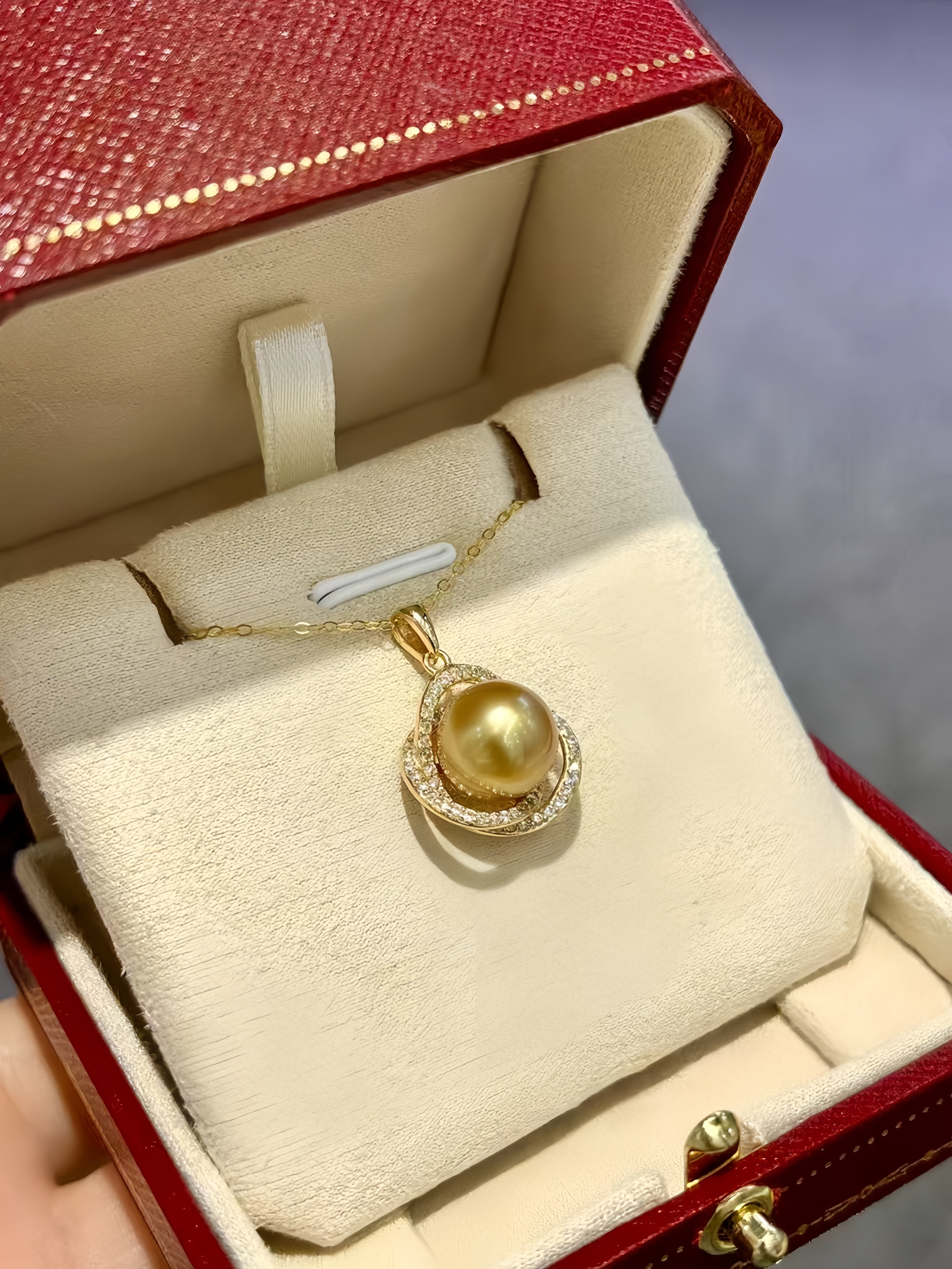 A gold pendant necklace with a large round gold pearl and small clear stones, displayed inside an open red jewelry box with cream velvet lining.