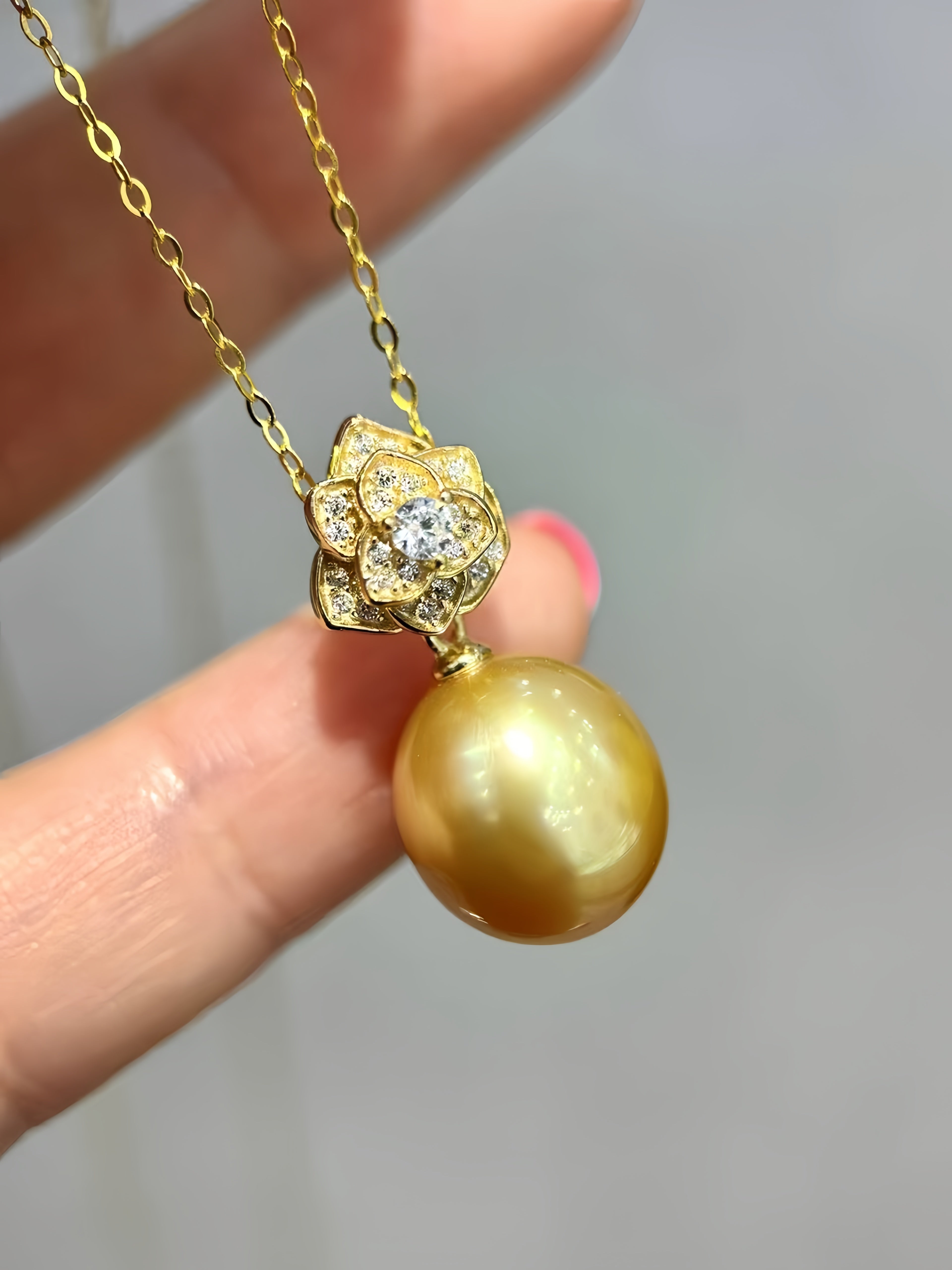 A gold pendant necklace featuring a large round gold pearl and a lotus-shaped setting with a central clear stone, held by a hand against a soft gray background.