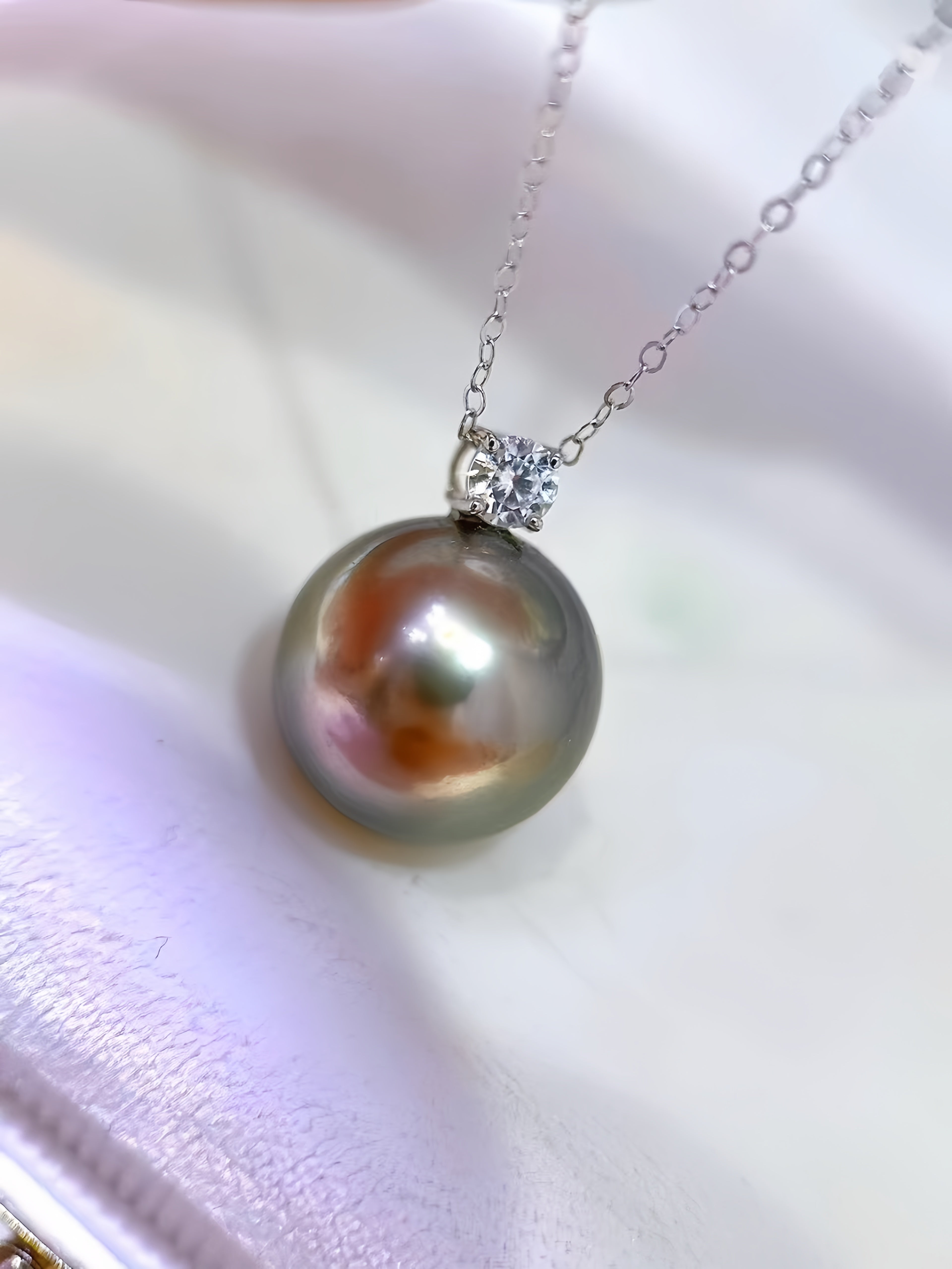 A close-up of a pearl pendant necklace on a delicate silver chain, resting on a soft white satin surface. The pearl has a pearlescent gray-green body with warm peach and orange iridescence, set in a silver bezel with a small clear accent stone.