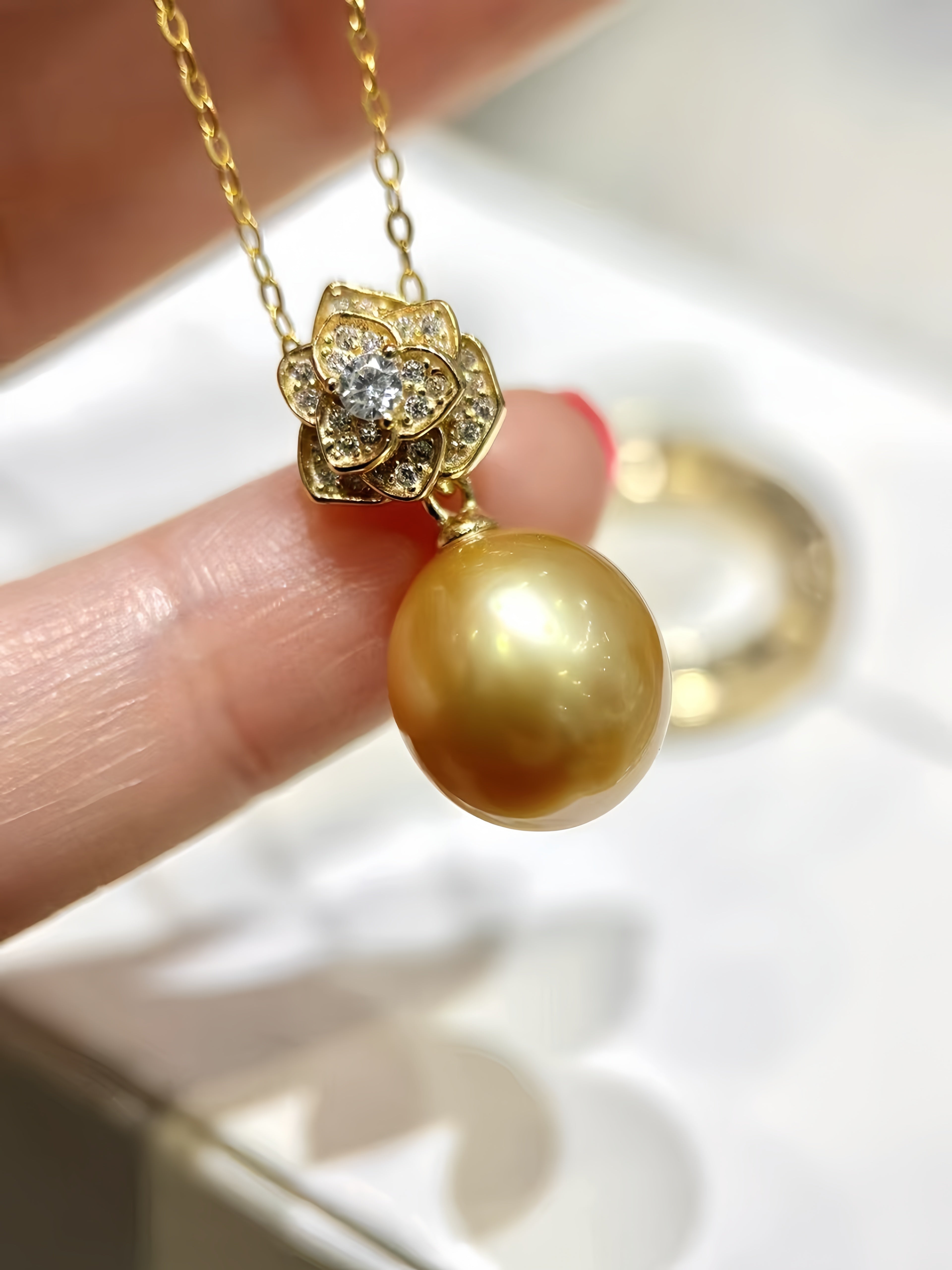A gold pendant necklace with a large round gold pearl and a flower-shaped setting with a clear center stone, held by a hand over a white jewelry box.