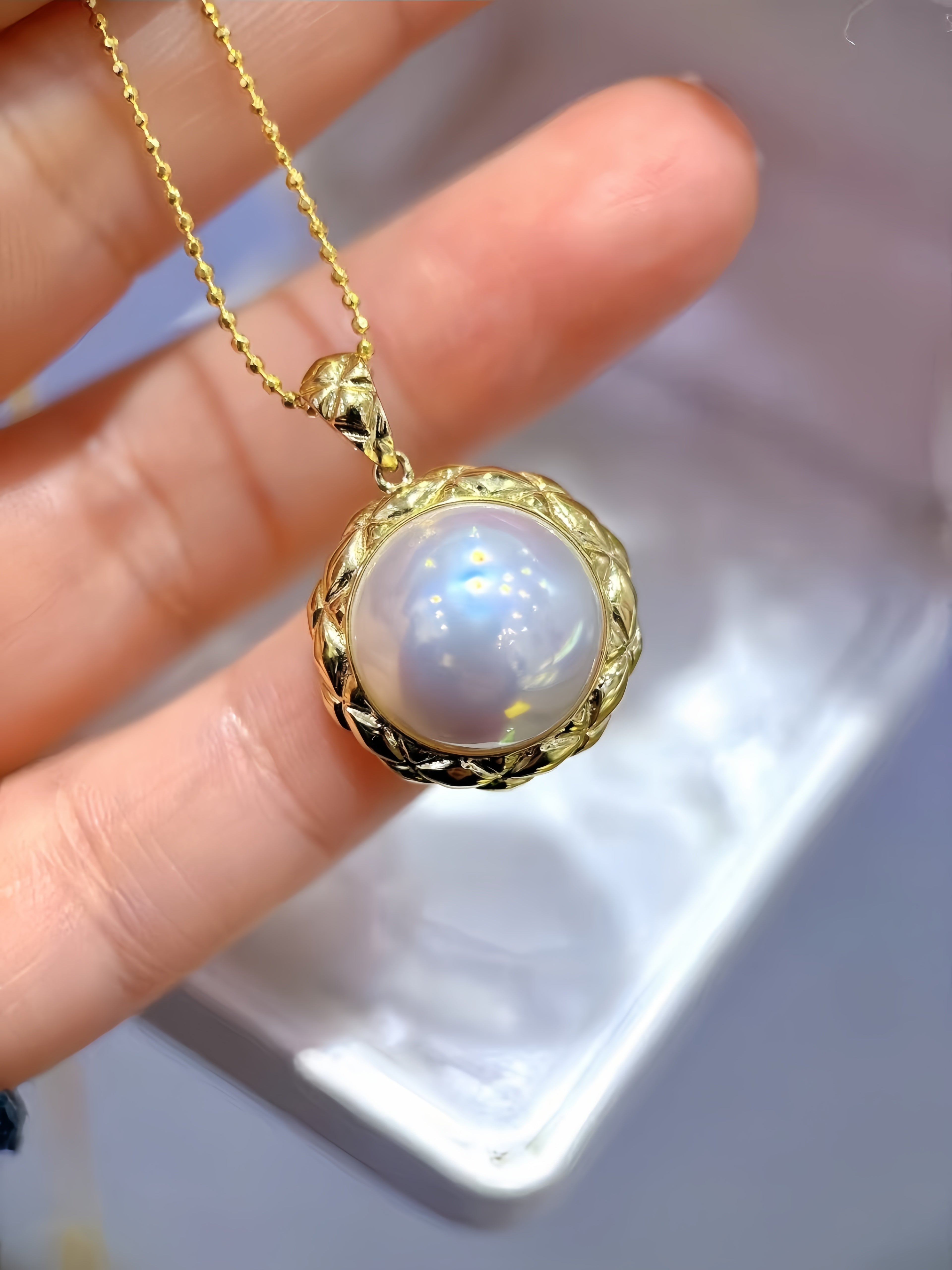 A gold pendant necklace with a round iridescent pearl-like center and a textured gold bezel, held in a person's hand against a soft white background.