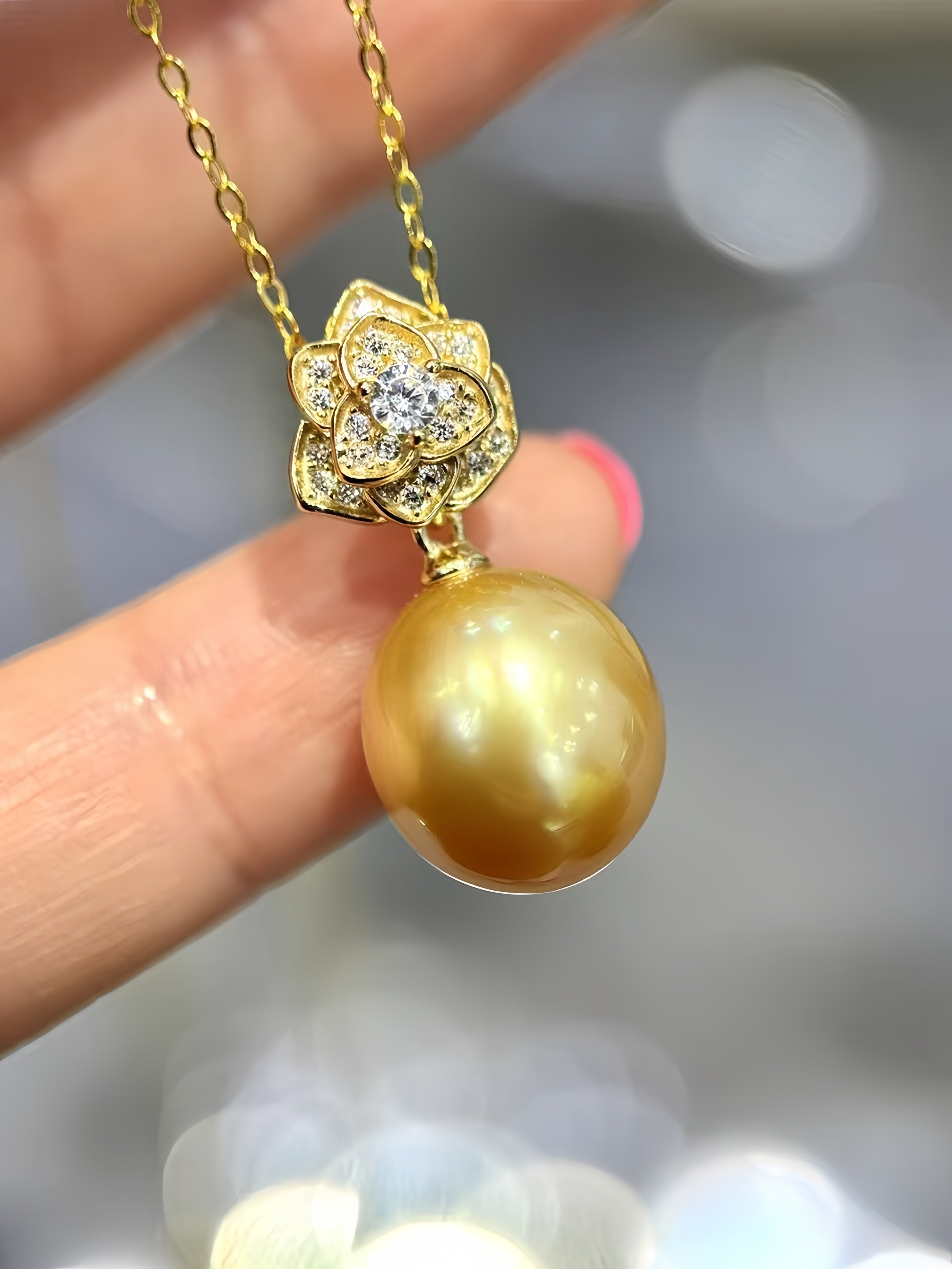 A gold pendant necklace featuring a large round gold pearl and a flower-shaped setting with small clear stones, held by a hand against a blurred gray background.