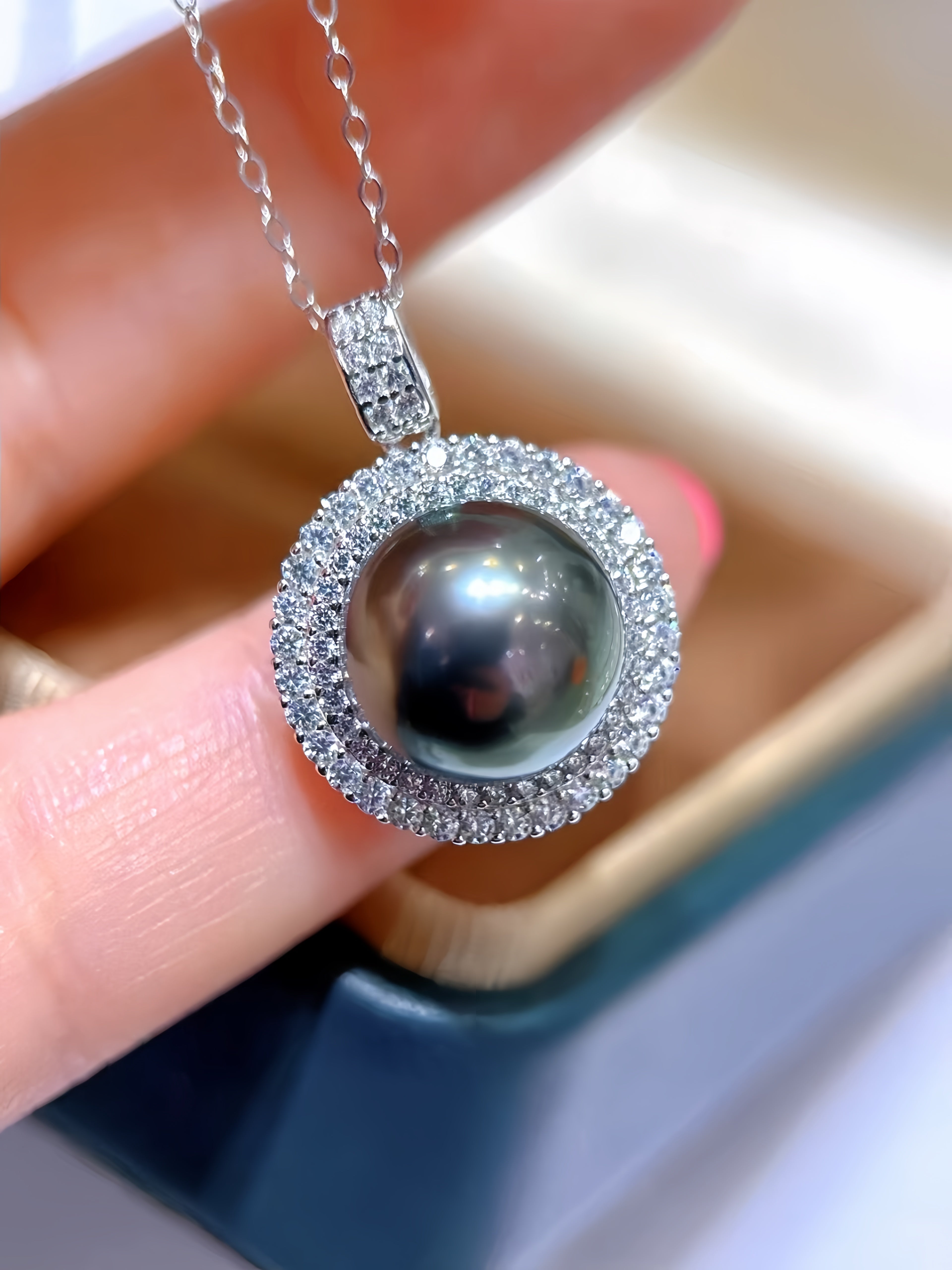 A close-up of a silver pendant necklace with a dark gray pearl surrounded by a halo of small clear stones, held by a hand over a jewelry box.