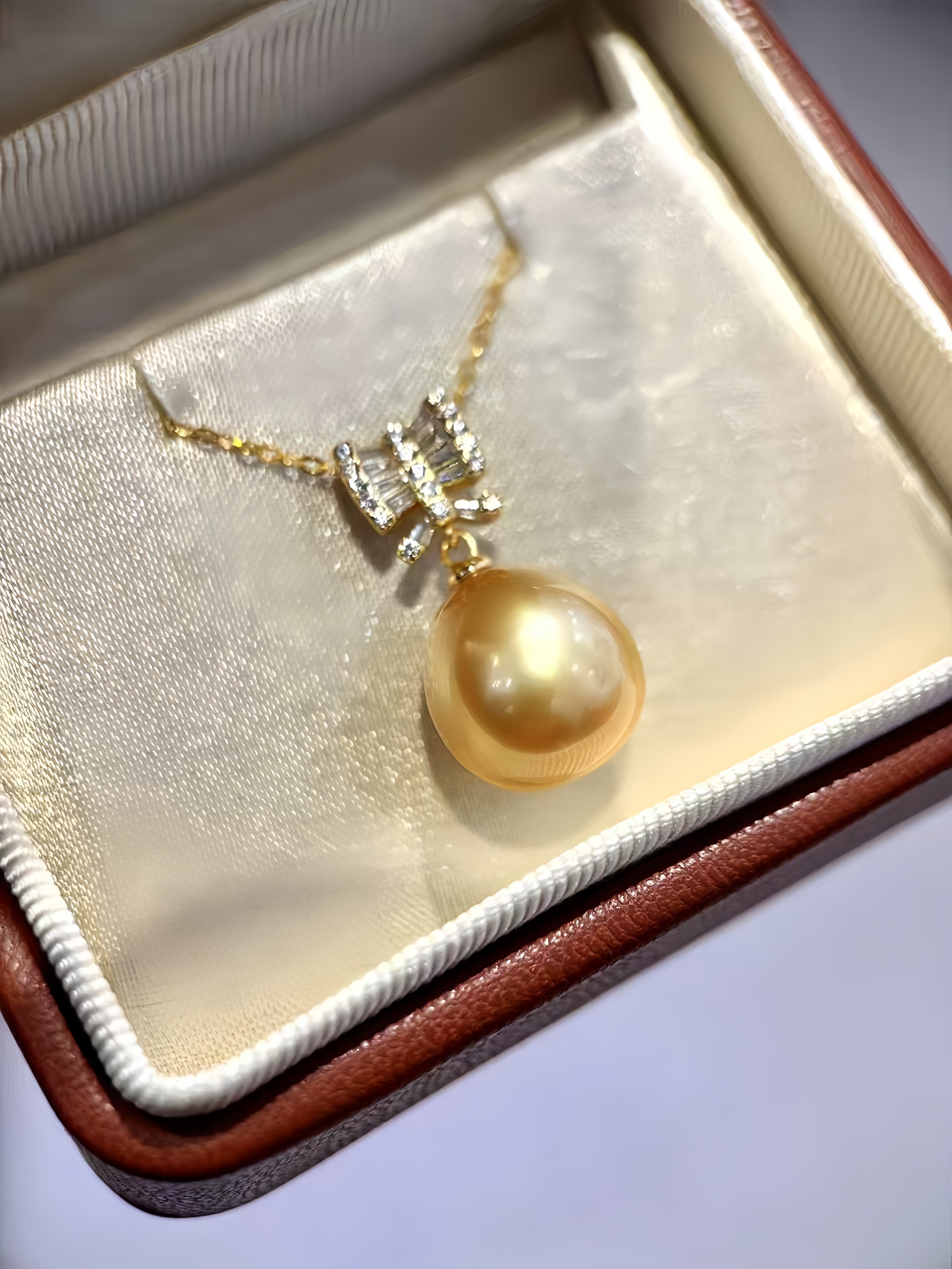 A gold pendant necklace with a large round yellow pearl and a small diamond-set bow detail, displayed inside an open brown jewelry box with cream satin lining.