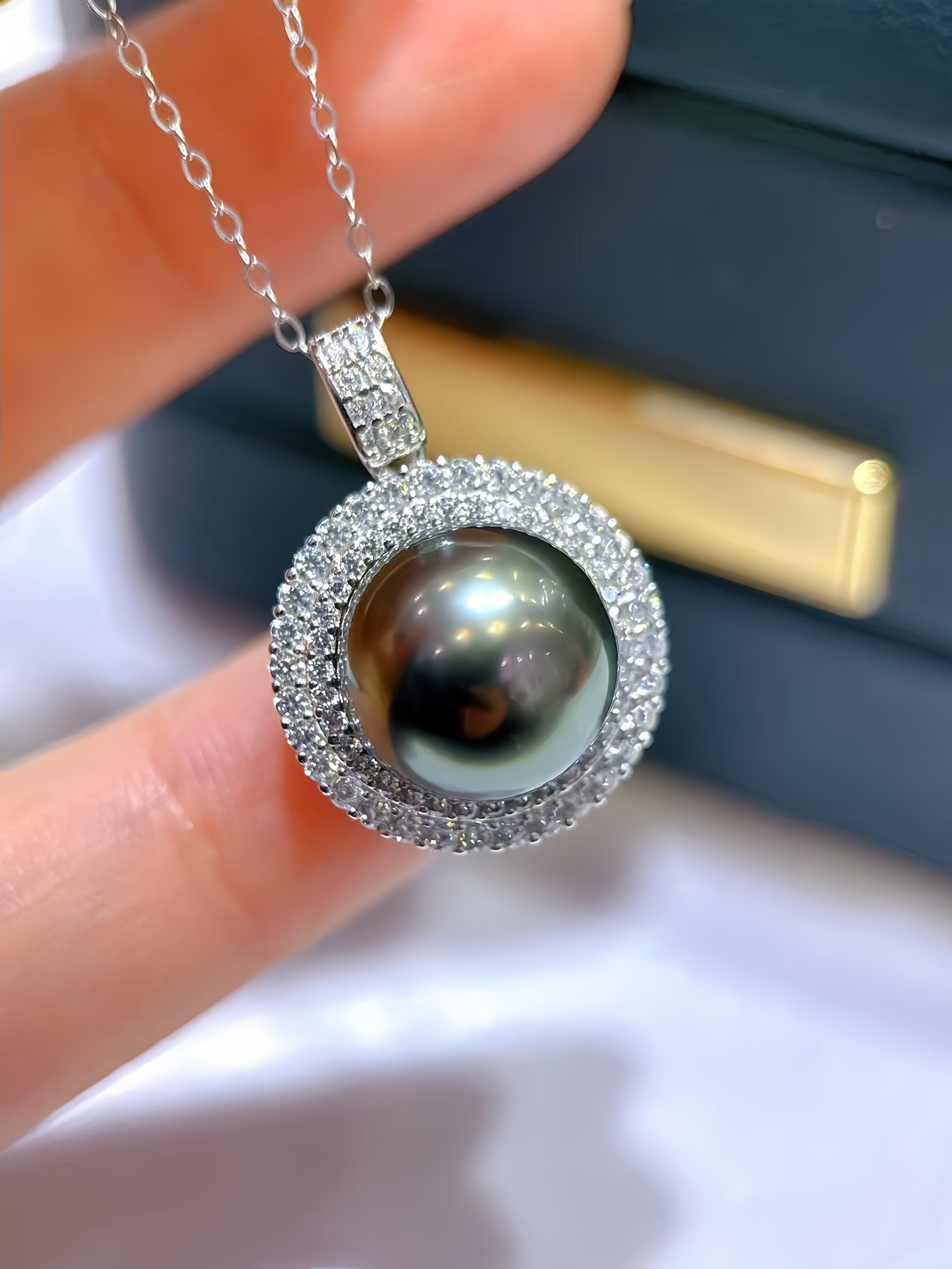A close-up of a silver pendant necklace with a large round black pearl surrounded by a halo of small clear stones, held between fingers with a dark jewelry box in the background.