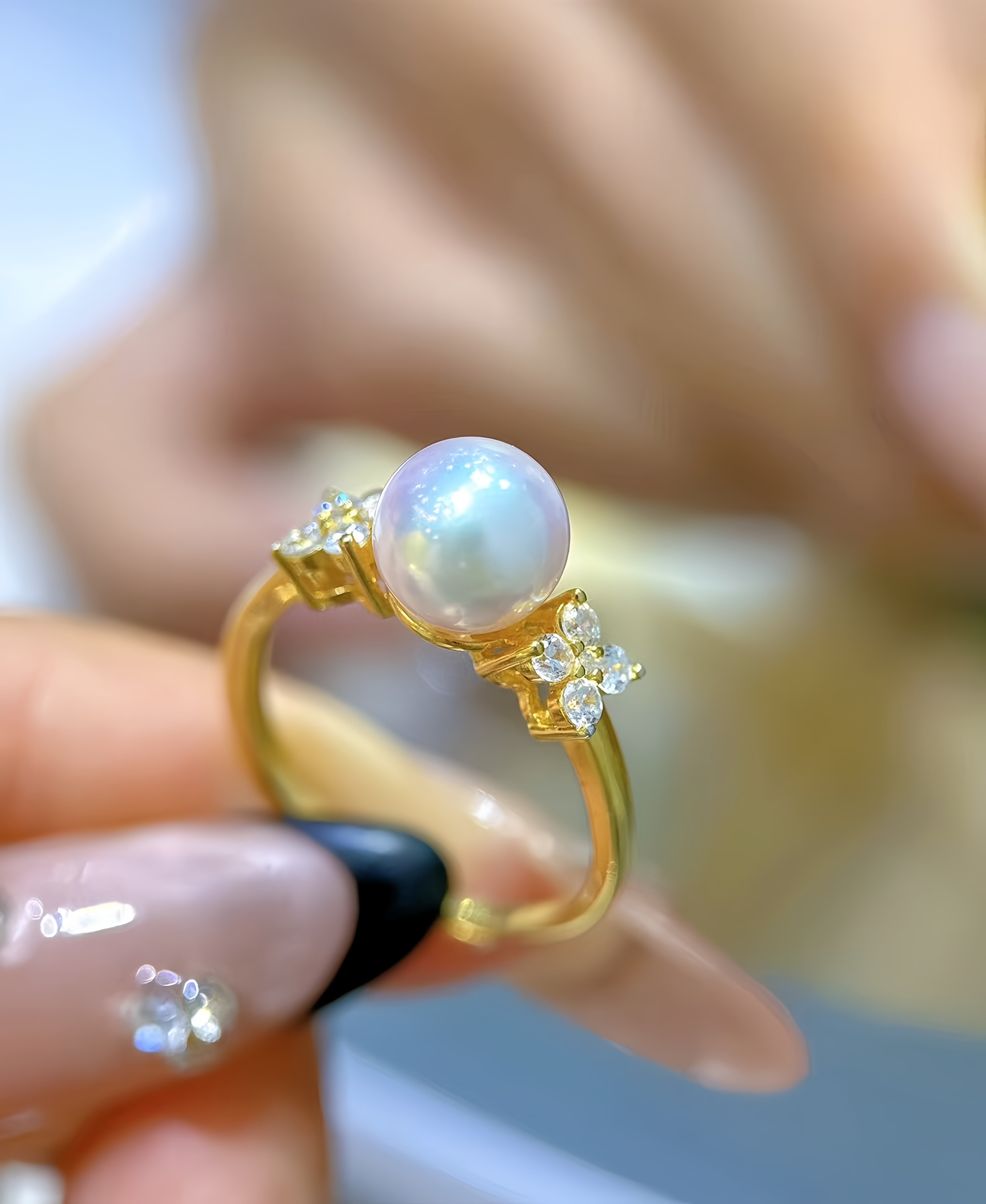 A gold ring with a round white pearl and small clear stones, held between fingers against a softly blurred background.