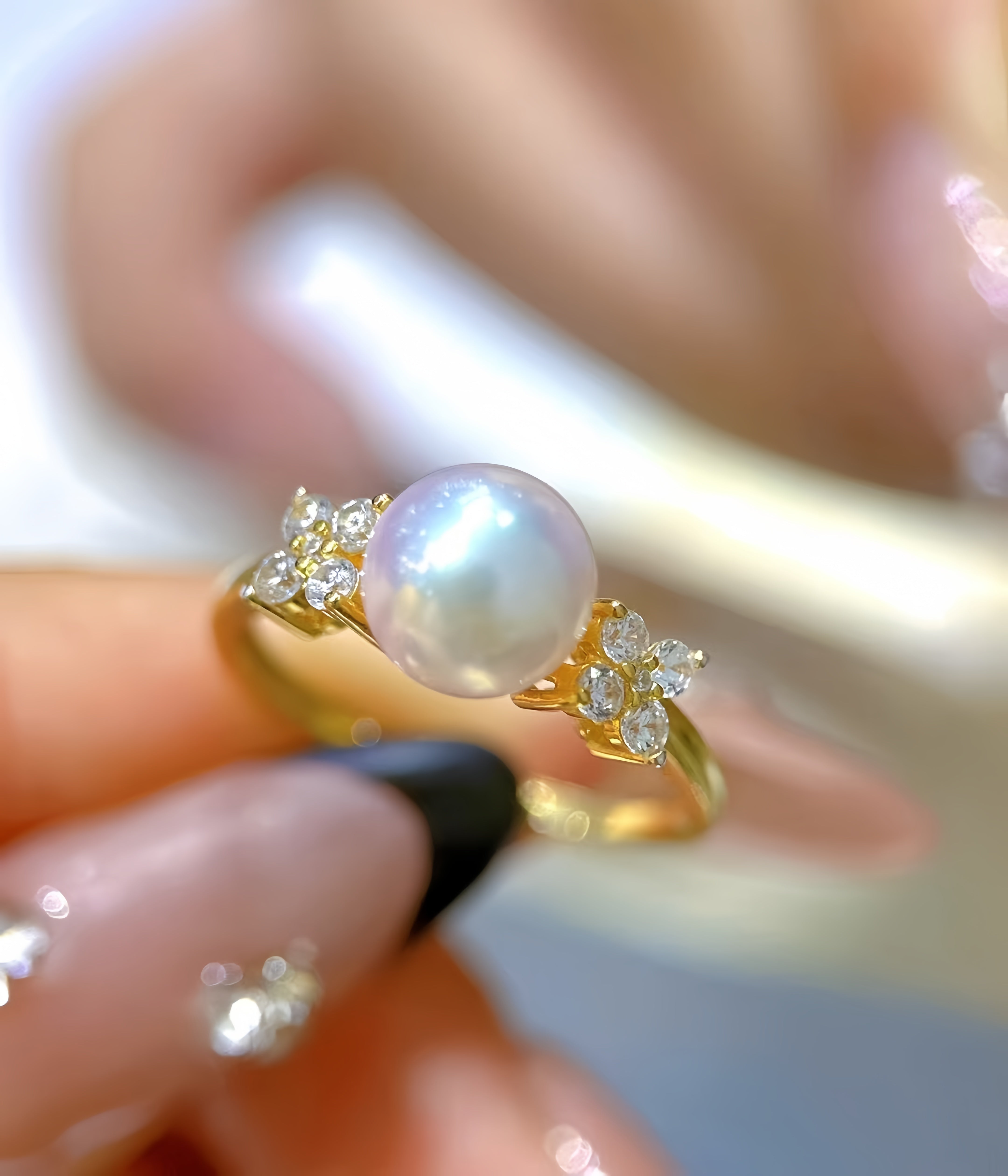 A gold ring with a large lustrous white pearl and small clear stones on either side, held between fingers against a soft blurred background.