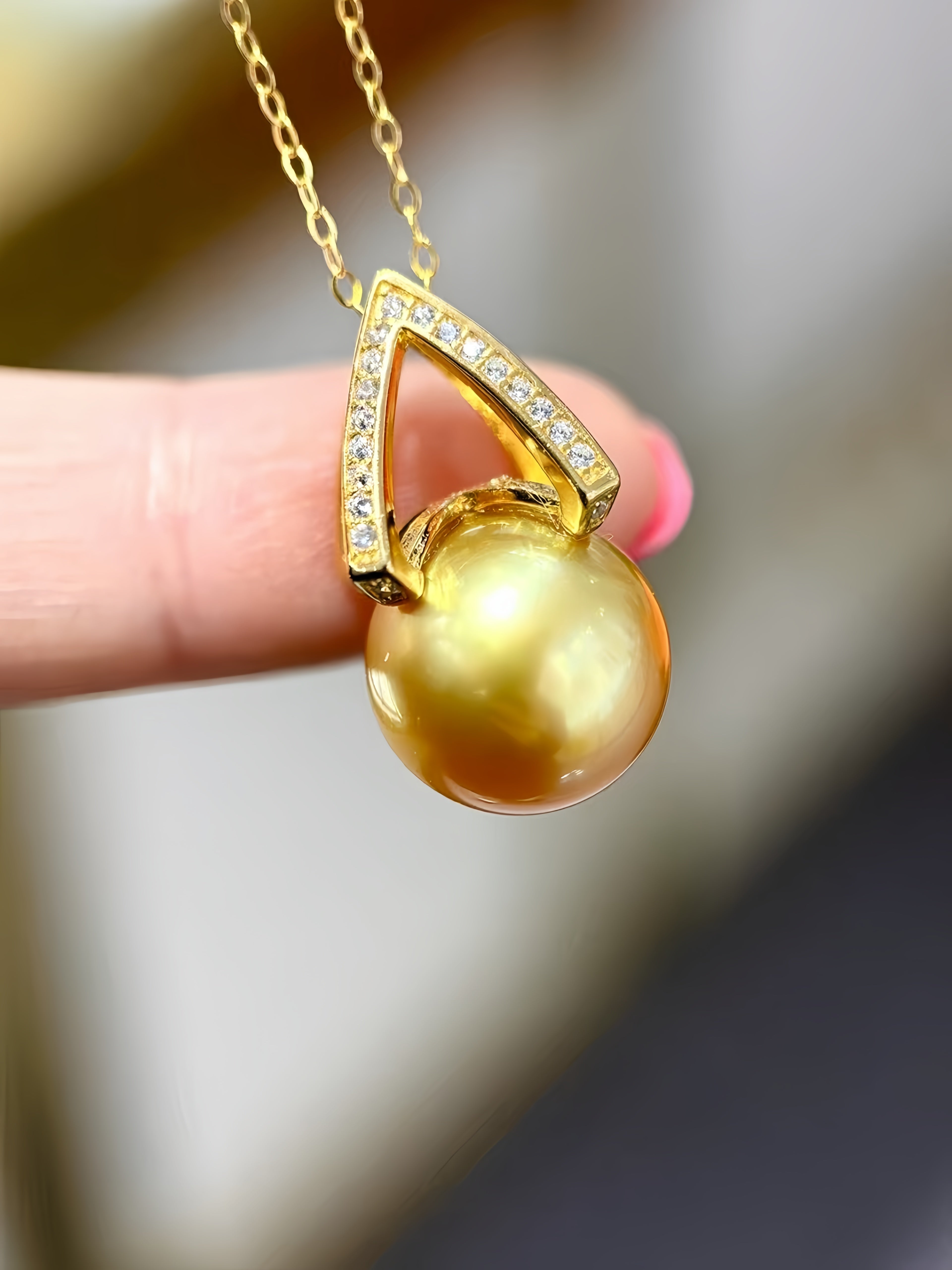 A gold pendant necklace featuring a large, lustrous gold pearl suspended beneath a geometric gold frame adorned with small clear stones, held by a person's fingers against a blurred background.