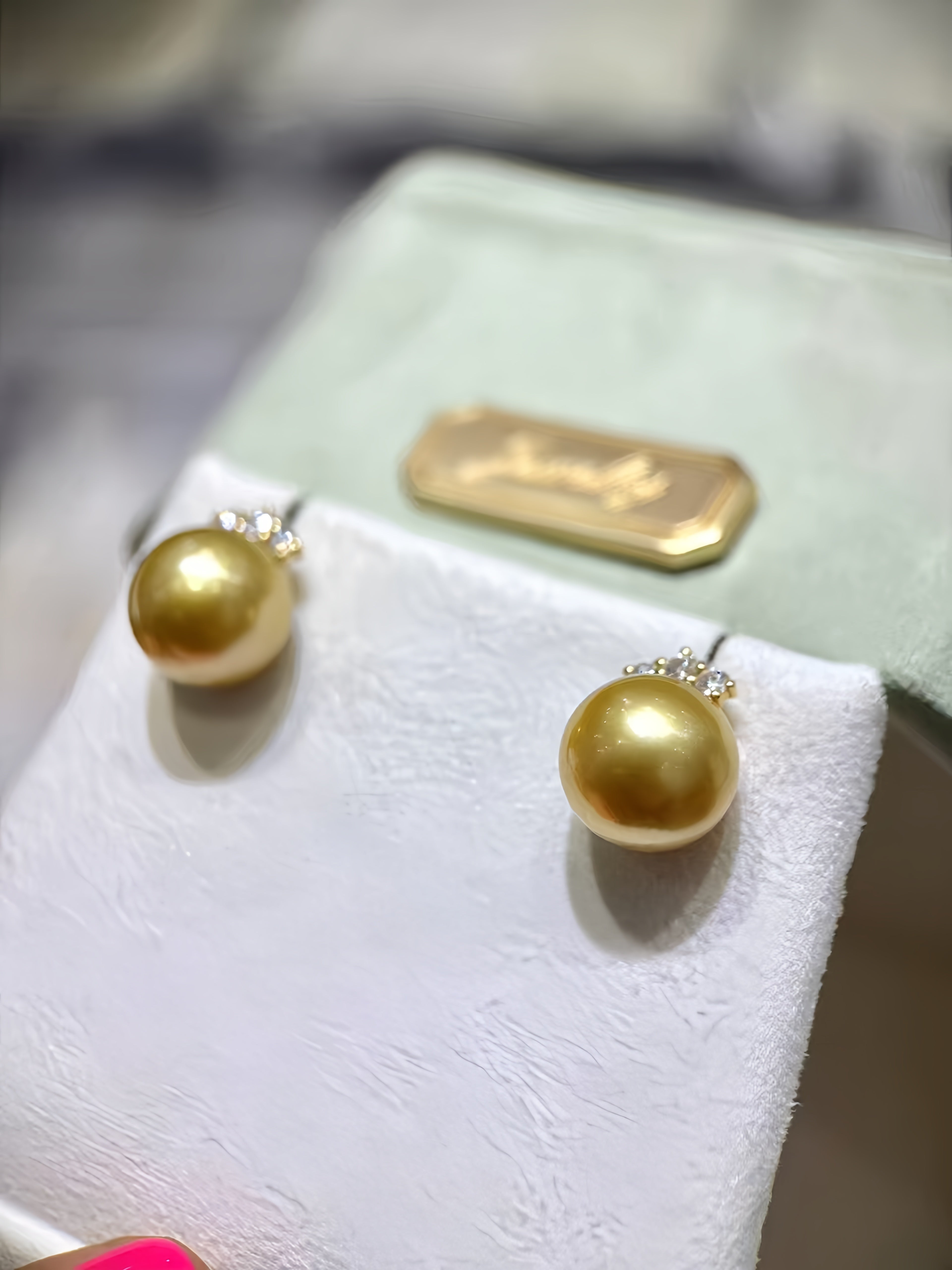 A pair of gold stud earrings with round gold pearls and small clear crystal accents, displayed on a white jewelry stand with a pale green box behind them.
