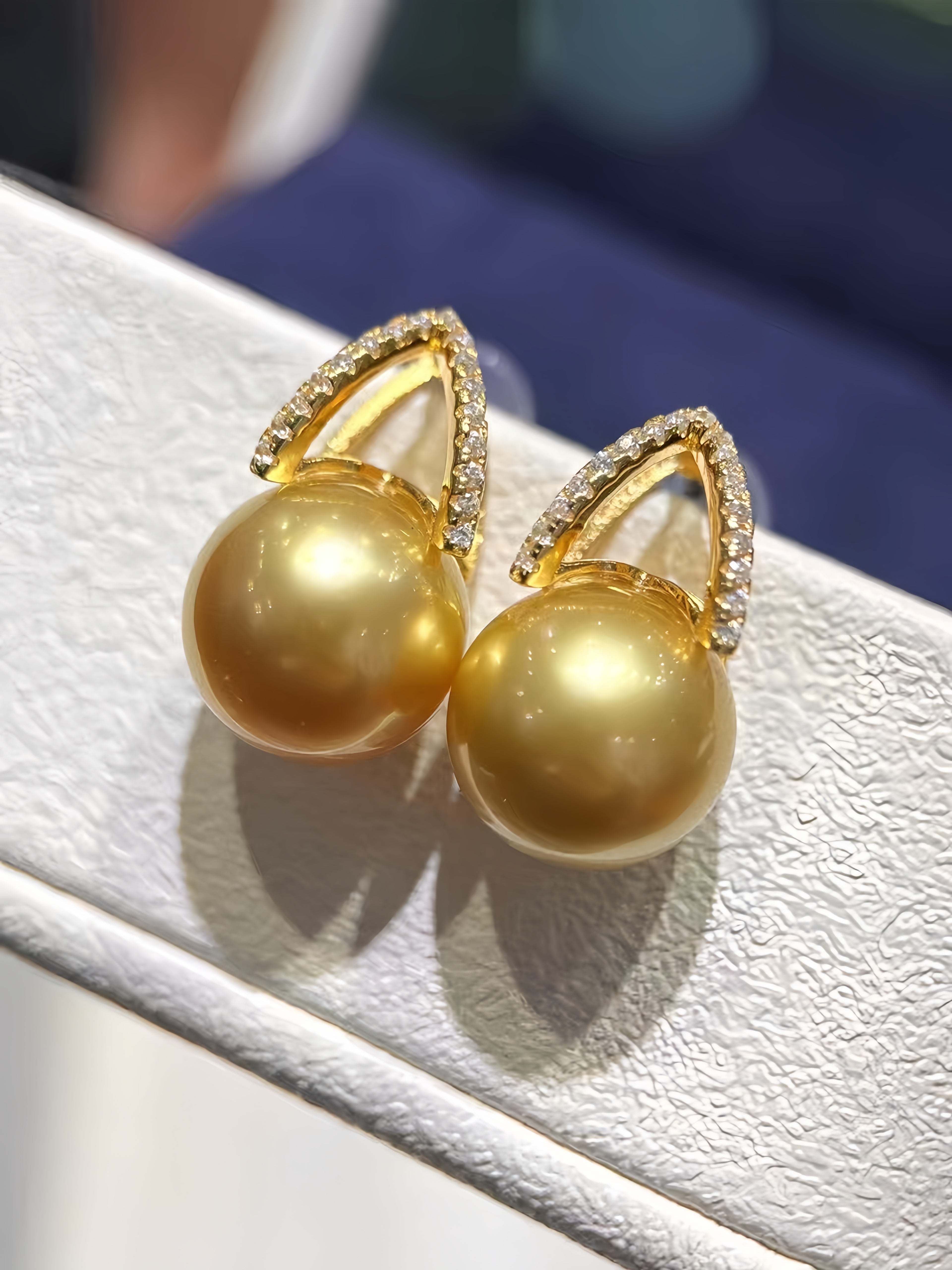 A pair of gold-toned drop earrings with large round pearl-like beads and small clear stone accents at the top, displayed on a textured white jewelry stand.