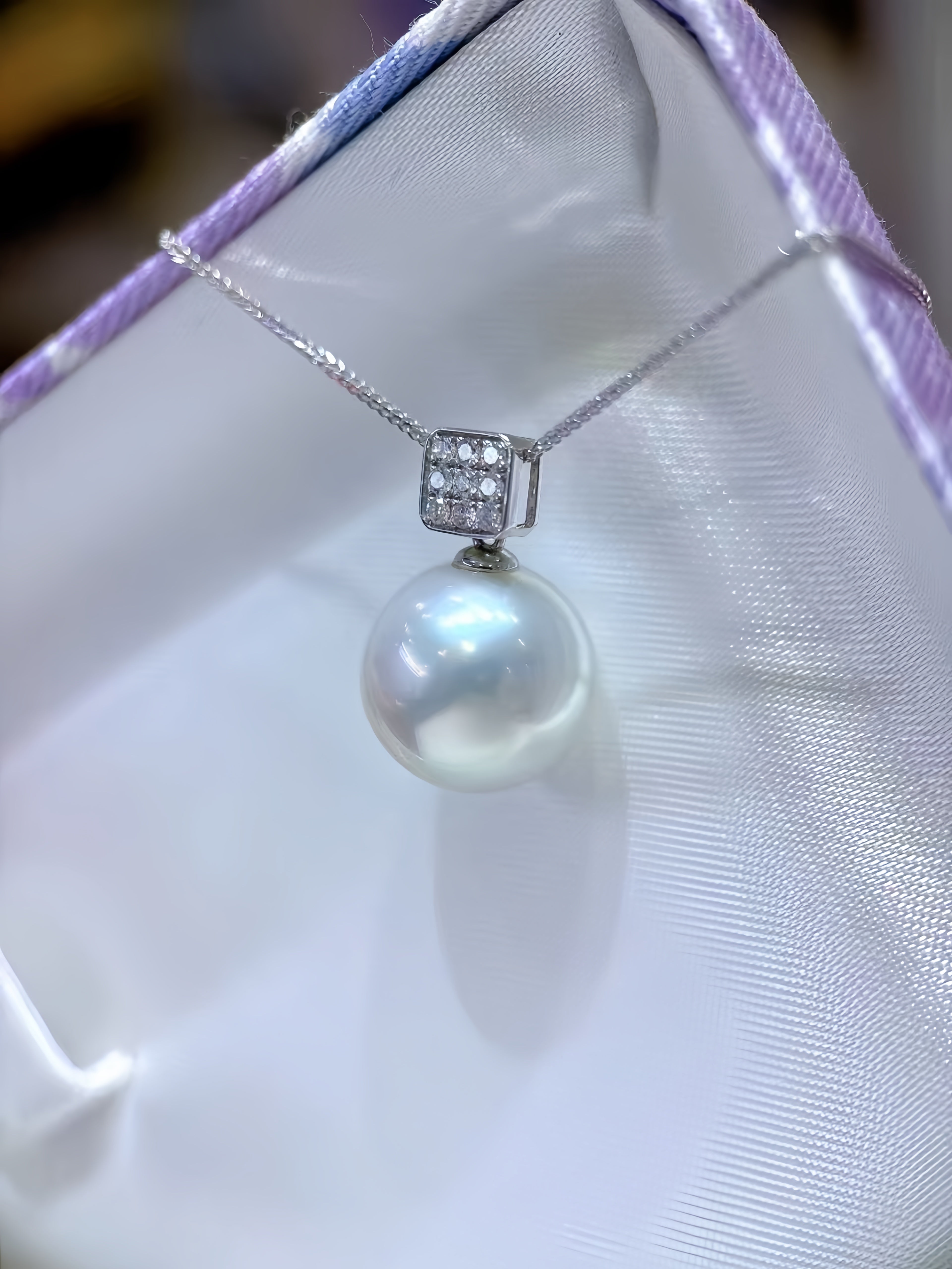 A silver pendant necklace featuring a single lustrous white pearl and a square bezel-set crystal accent, displayed on a white satin surface.