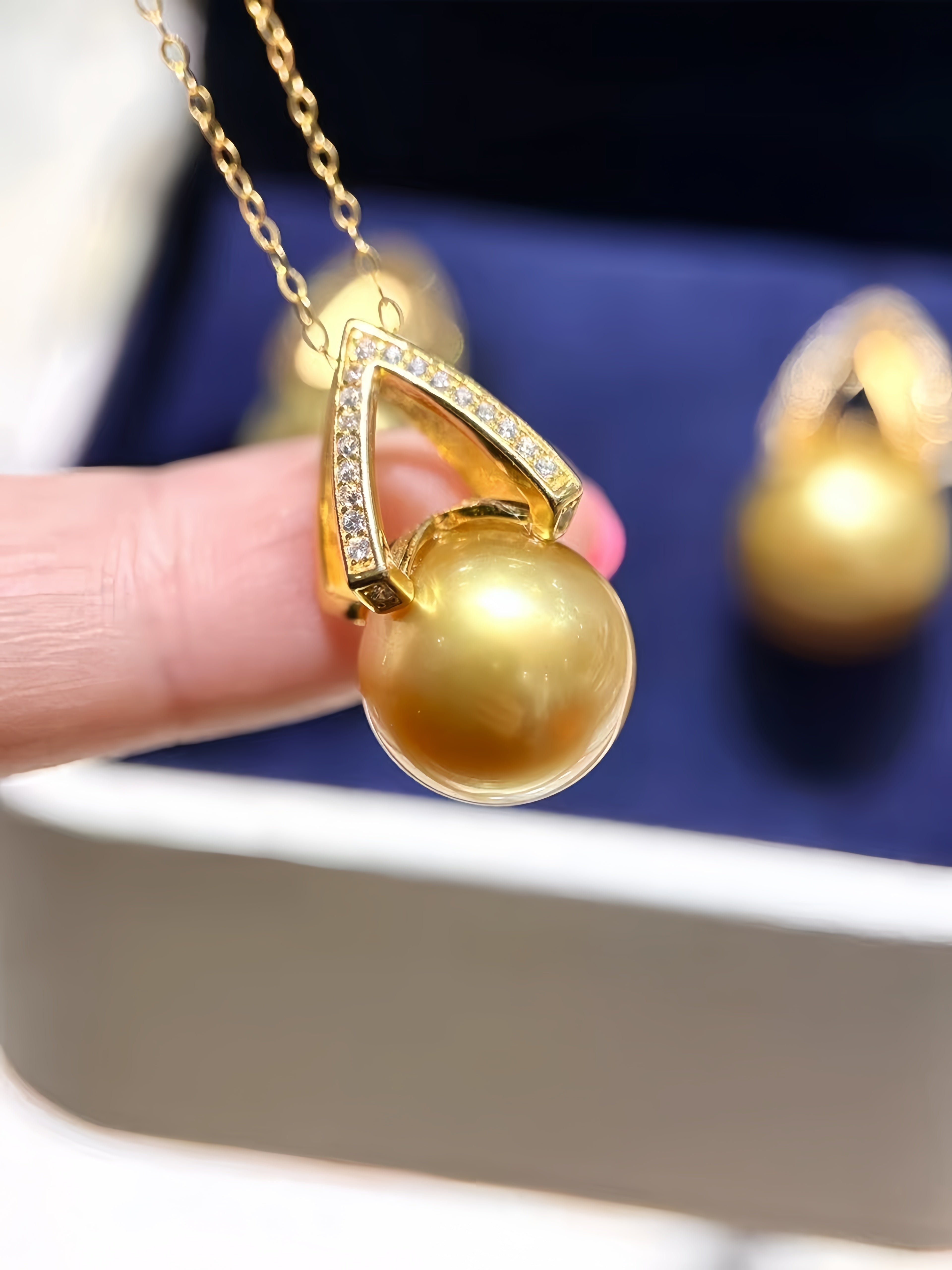 A gold pendant necklace with a large round pearl and a diamond-accented triangular frame, held by a hand in front of a blue jewelry box.