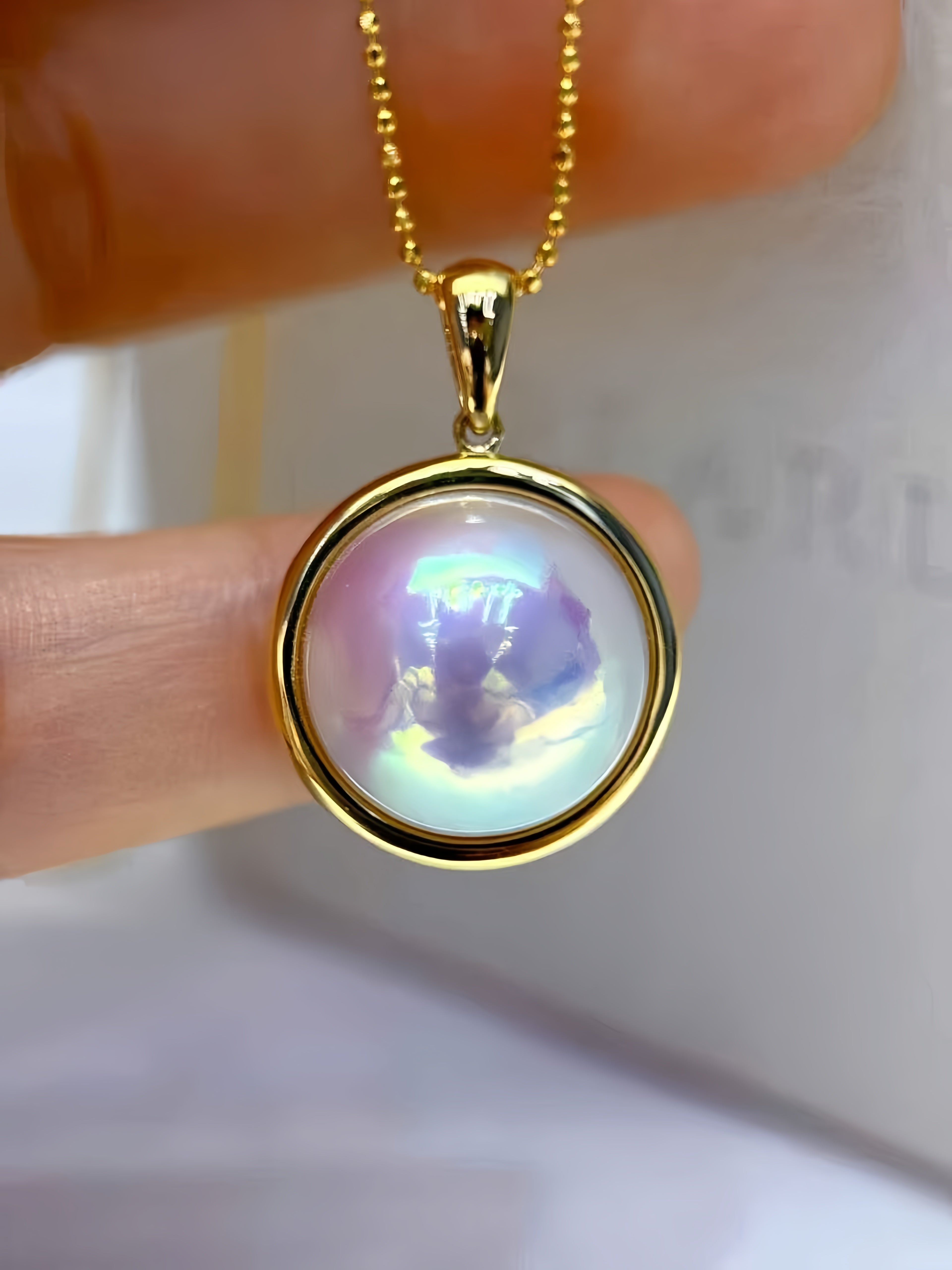 A gold pendant necklace with a round iridescent moonstone or pearl-like cabochon, held between fingers against a soft white background.