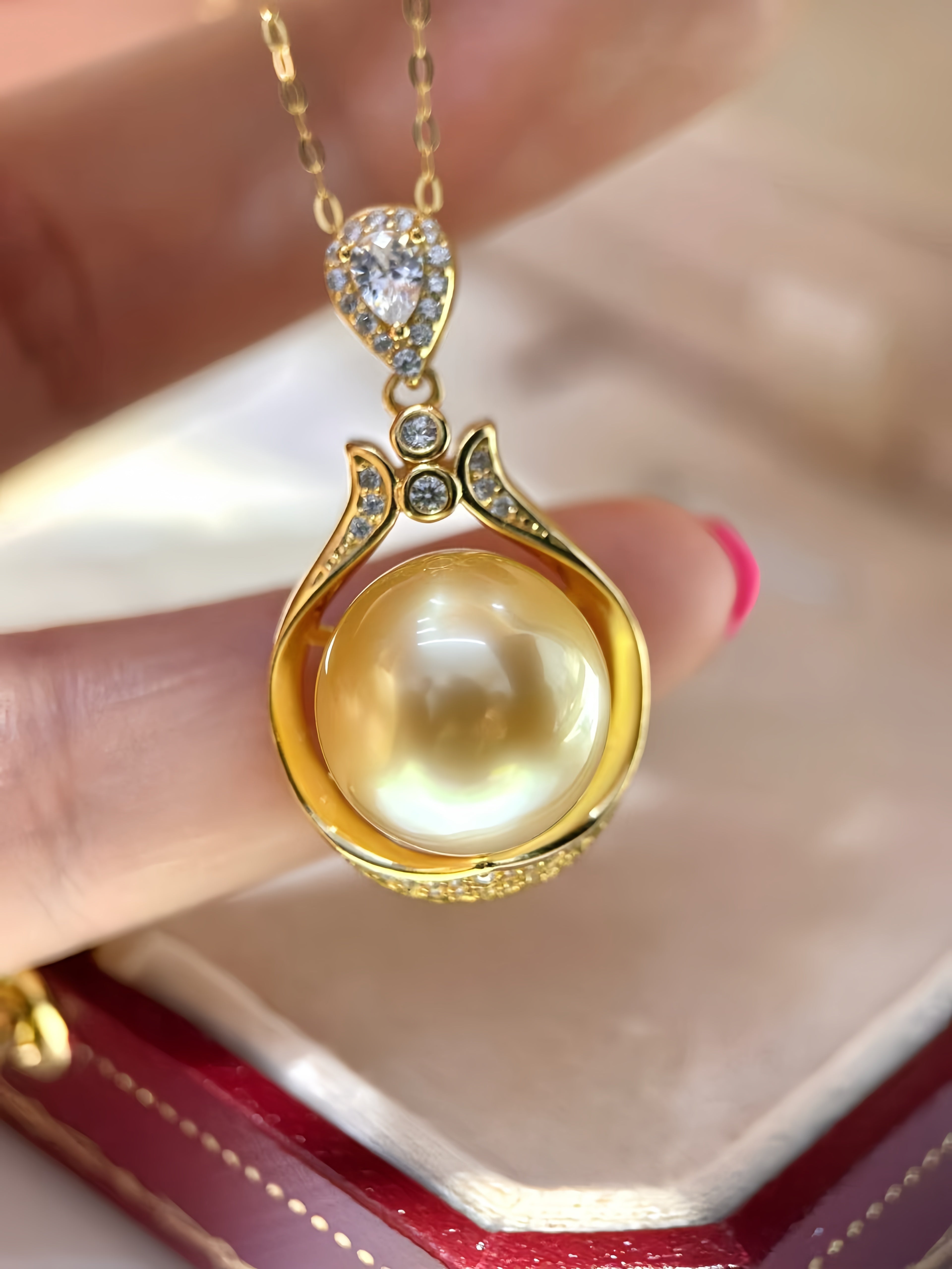 A gold pendant necklace with a large round yellow pearl and small clear stones, held between fingers above an open red jewelry box.