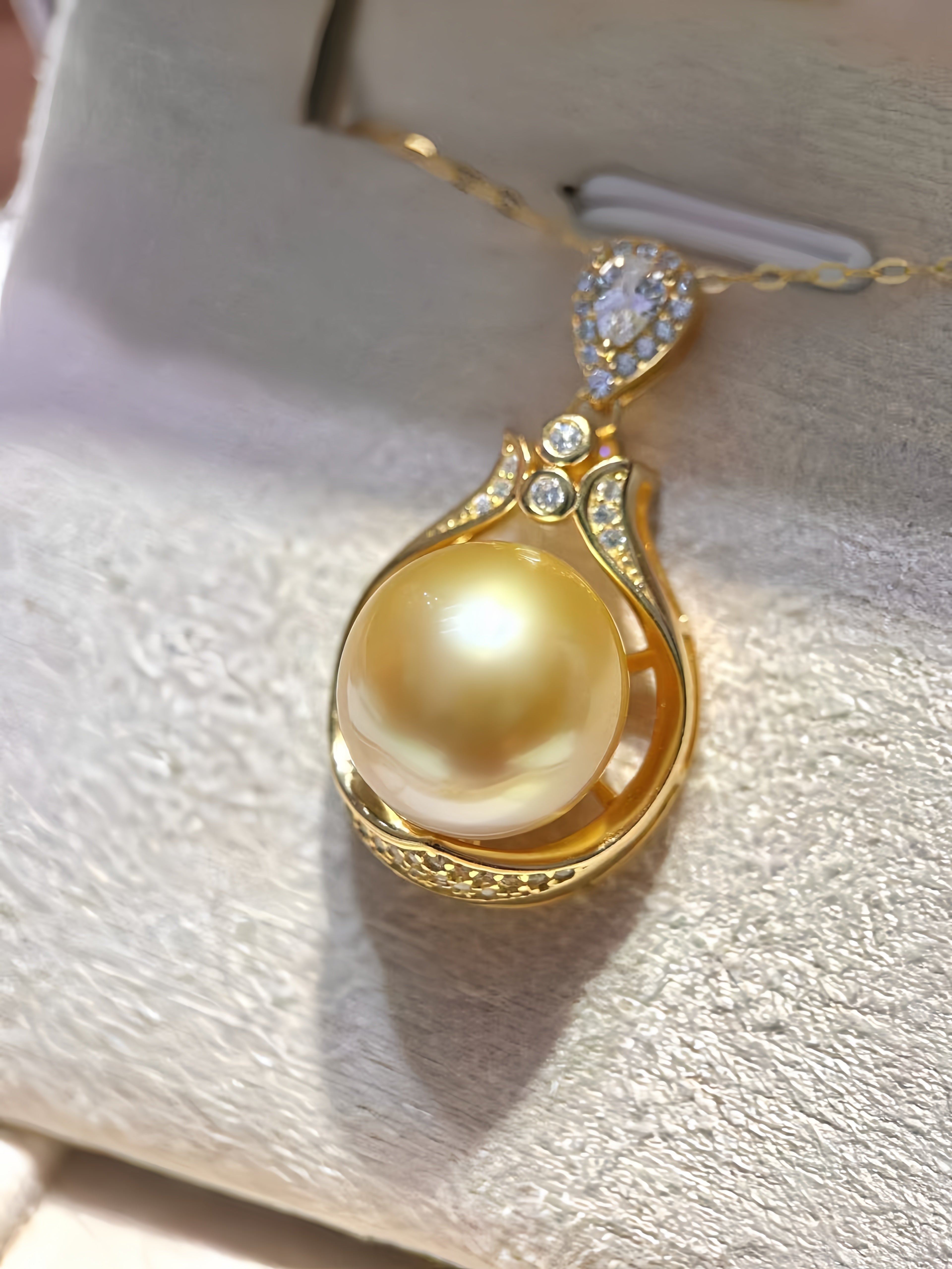 A gold pendant necklace featuring a large round yellow pearl in a gold teardrop-shaped setting, accented with small clear stones, displayed on a textured light beige jewelry box.