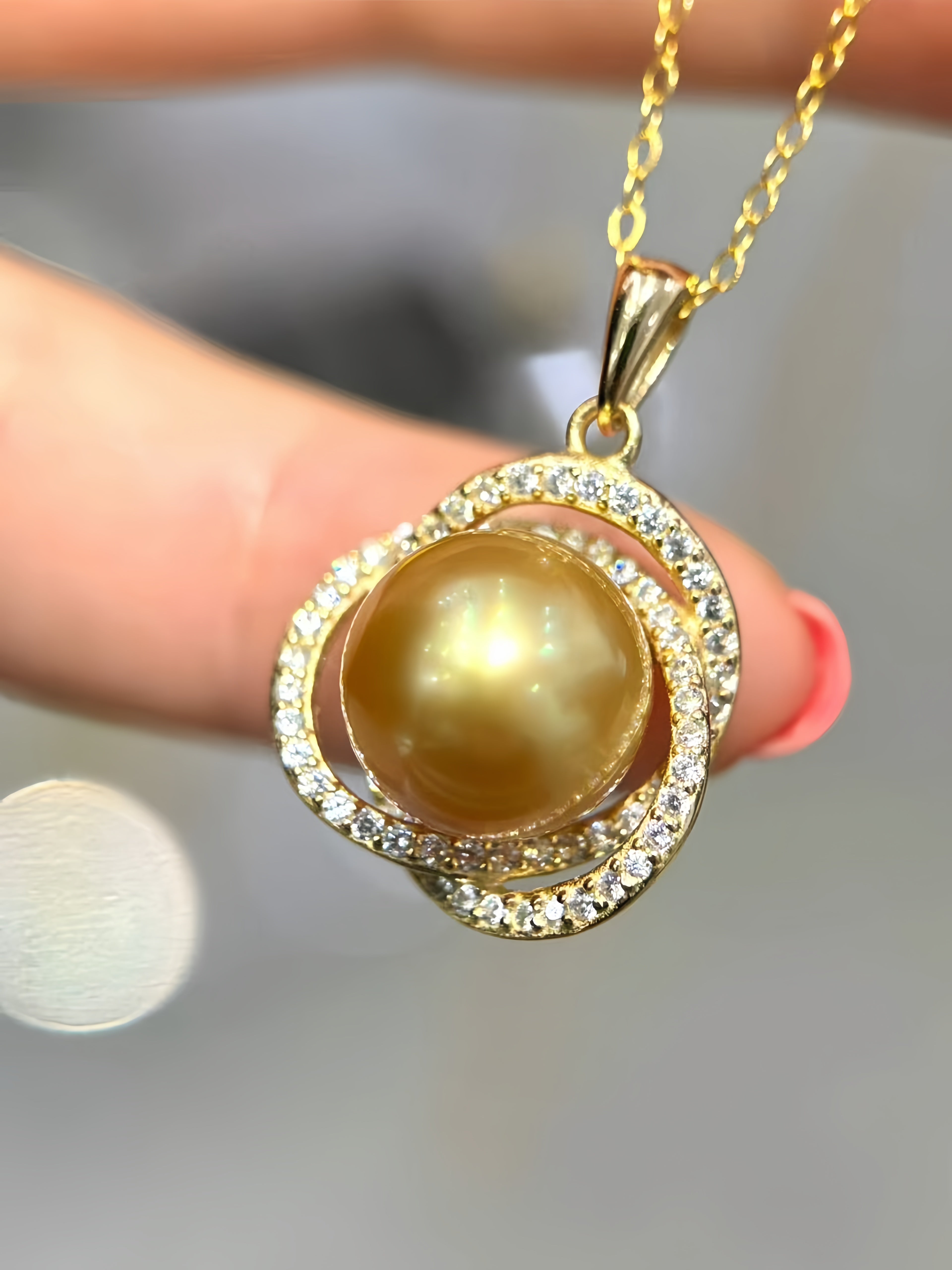 A gold pendant necklace featuring a large round golden pearl surrounded by double halos of small clear crystals, held between fingers against a soft gray background.