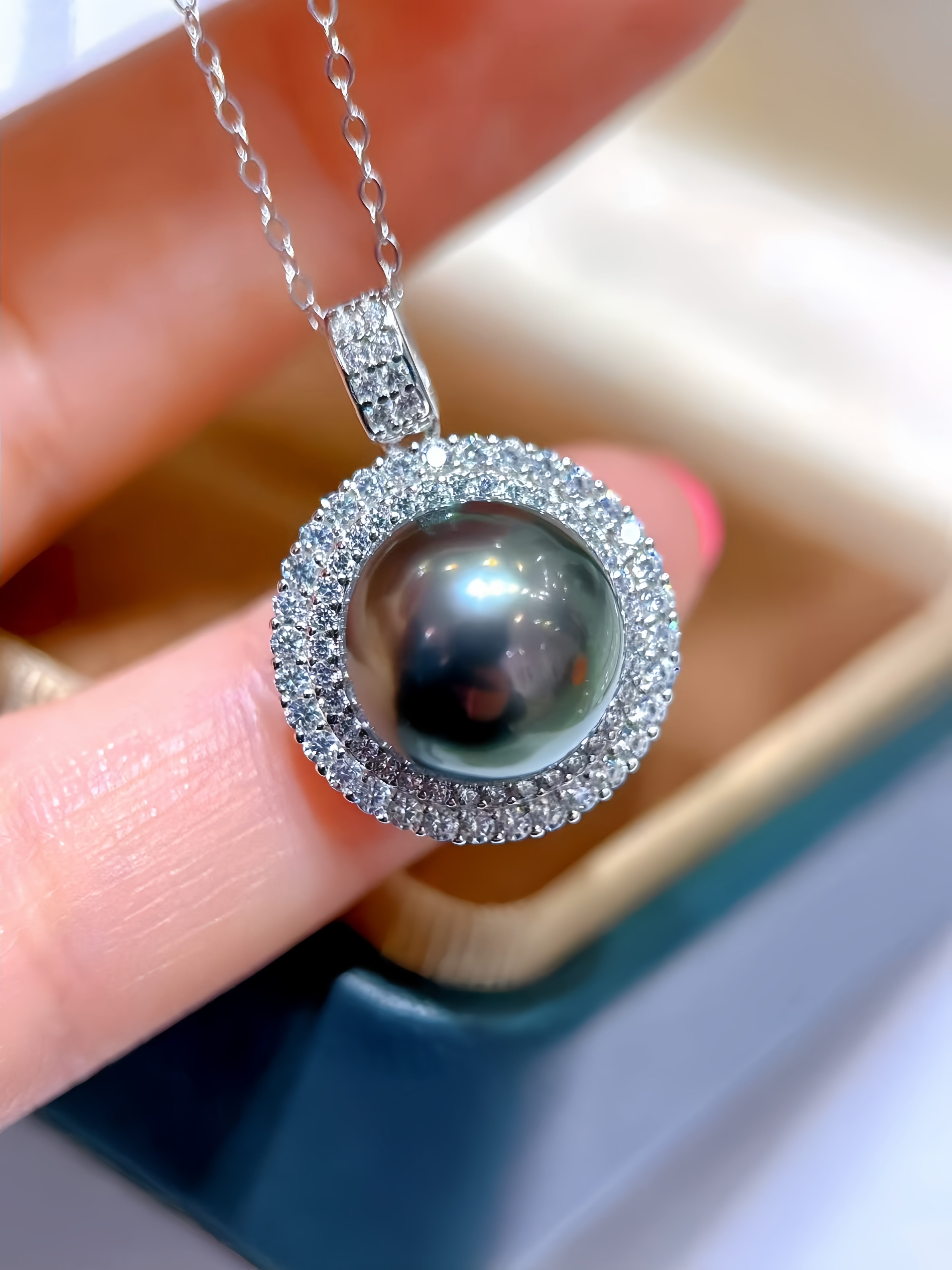 A close-up of a silver pendant necklace with a dark pearl surrounded by a halo of small clear stones, held between fingers with a jewelry box in the background.