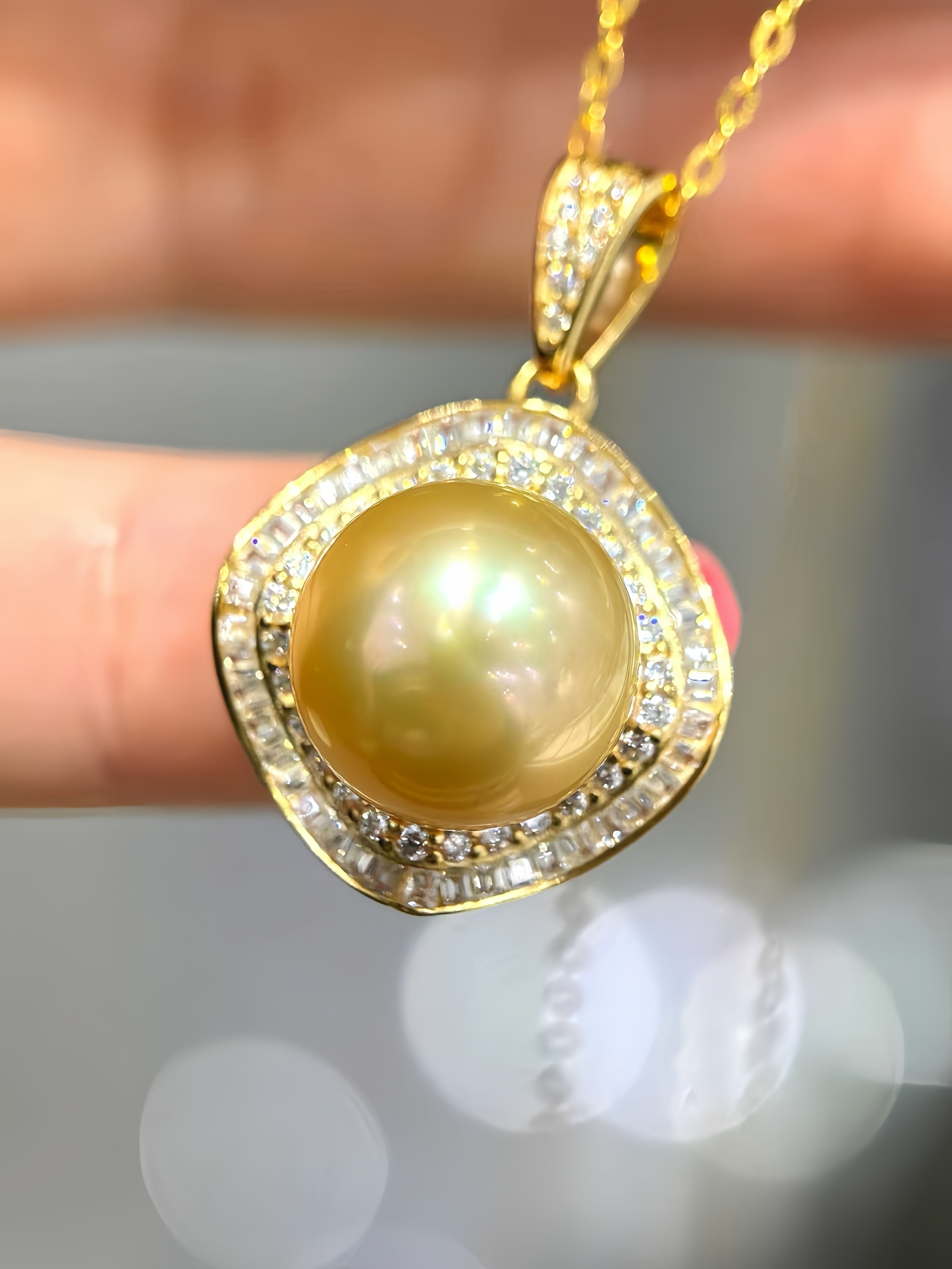 A gold pendant necklace featuring a large round yellow pearl surrounded by a halo of small clear stones, held between fingers against a blurred gray background.