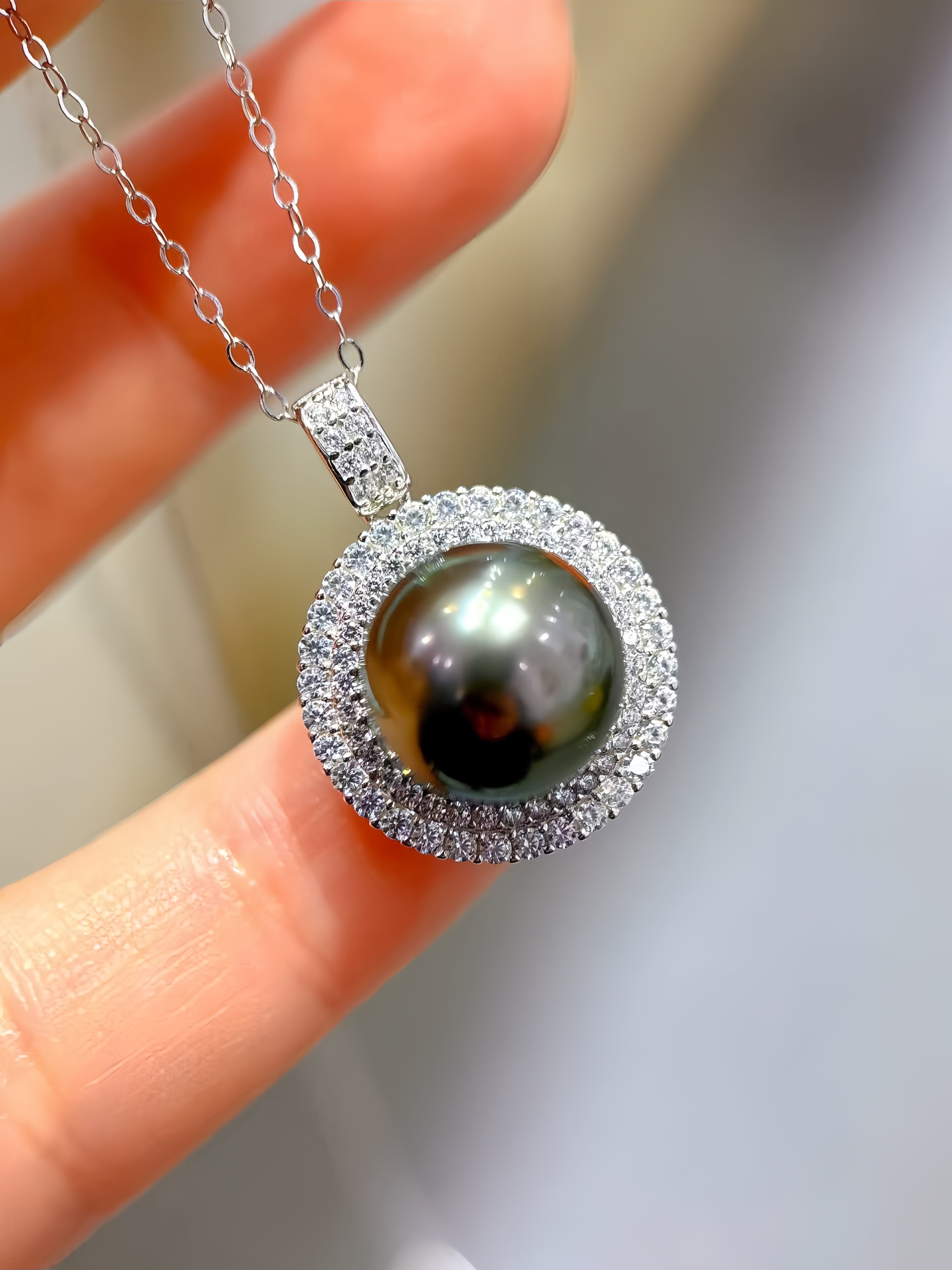 A close-up of a silver pendant necklace held between fingers. The pendant features a large, round, dark pearl surrounded by a double halo of small clear stones.