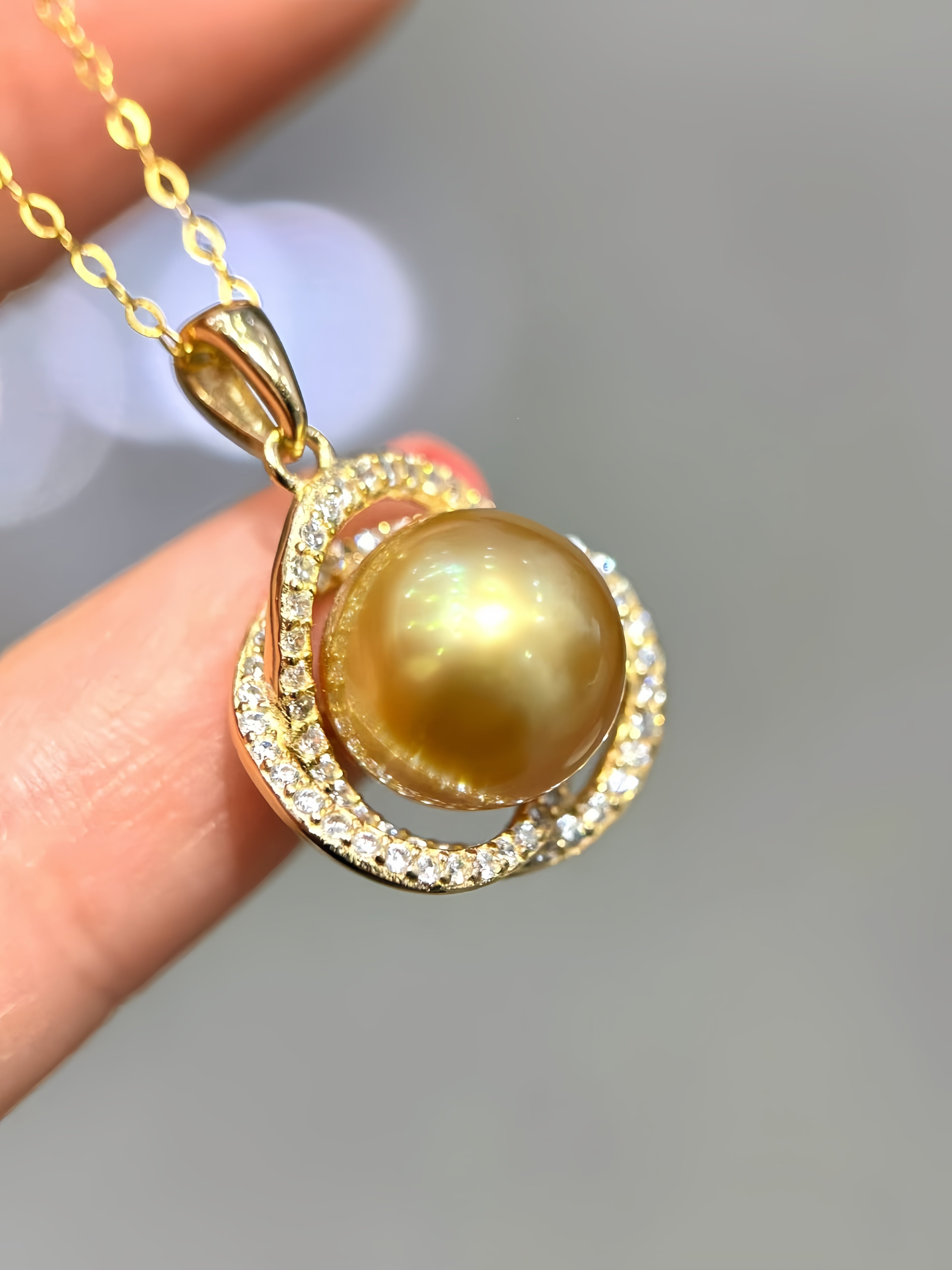 A gold pendant necklace with a large iridescent golden pearl surrounded by double rings of small clear stones, held between fingers against a soft gray background.