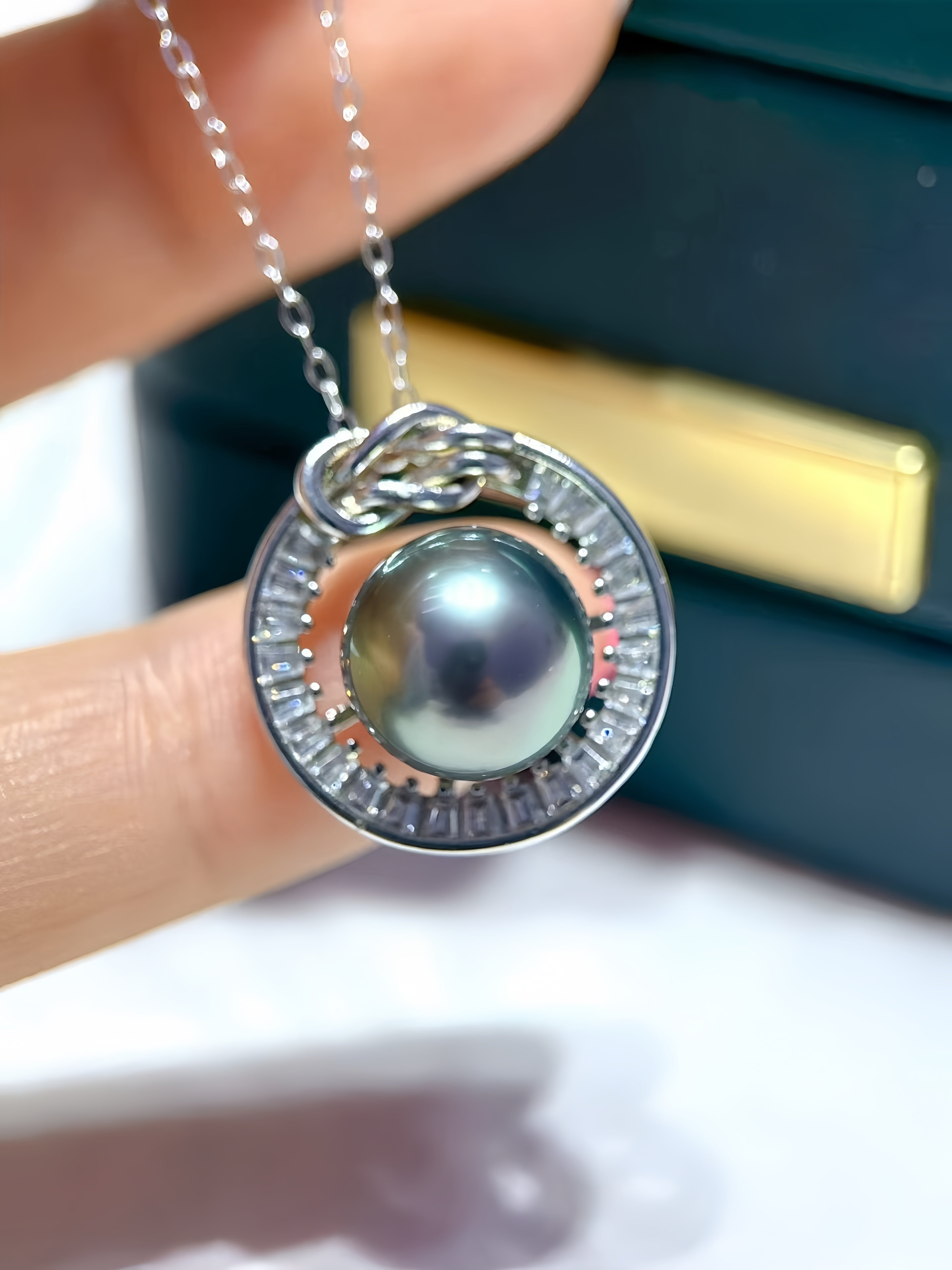 A close-up of a silver pendant necklace with a dark gray pearl and clear stones, held by a person's fingers in front of a dark jewelry box.