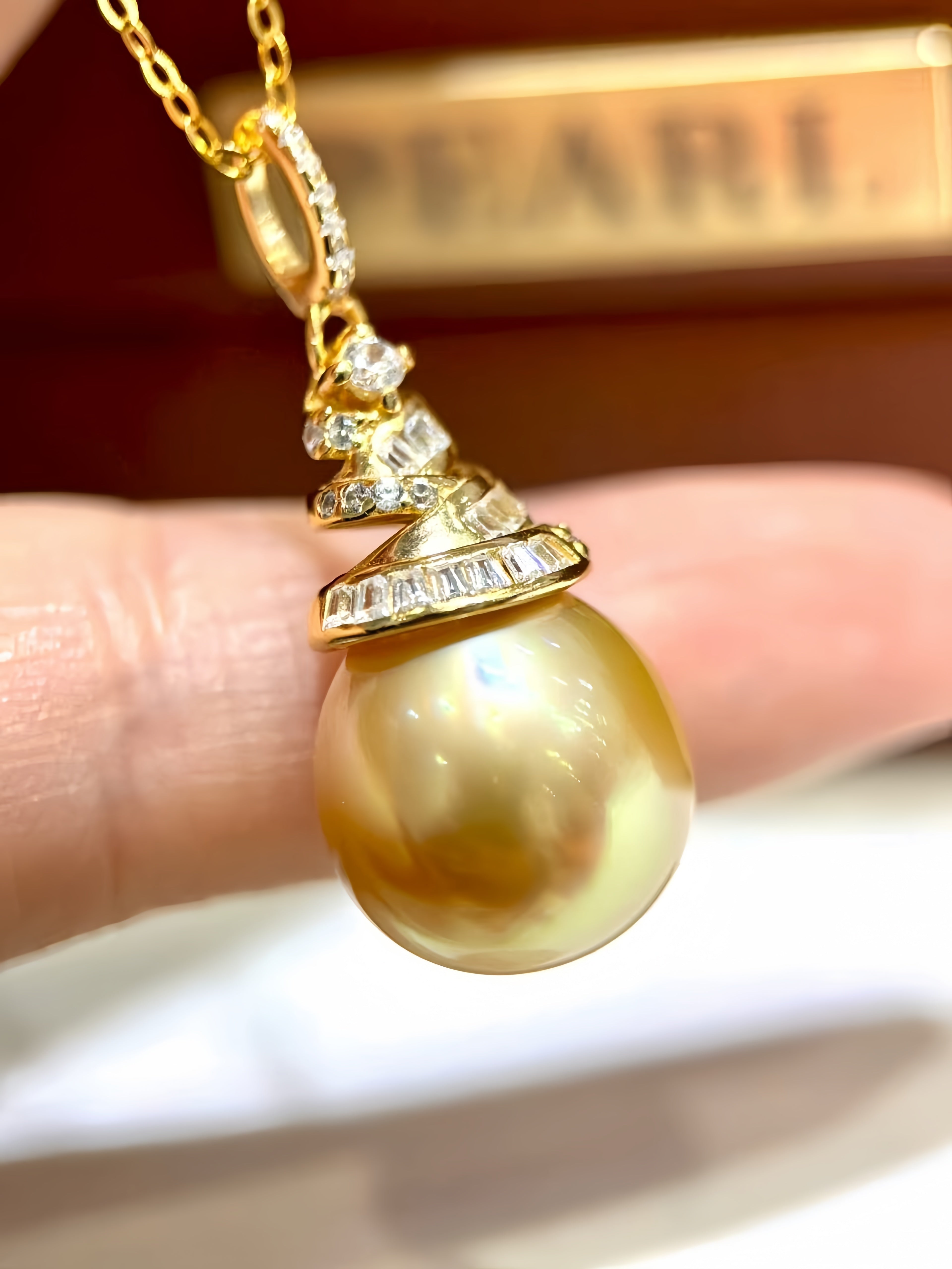 A gold pendant necklace featuring a large, lustrous pearl-like bead with a tiered gold and diamond crown detail, held by a person's fingers.
