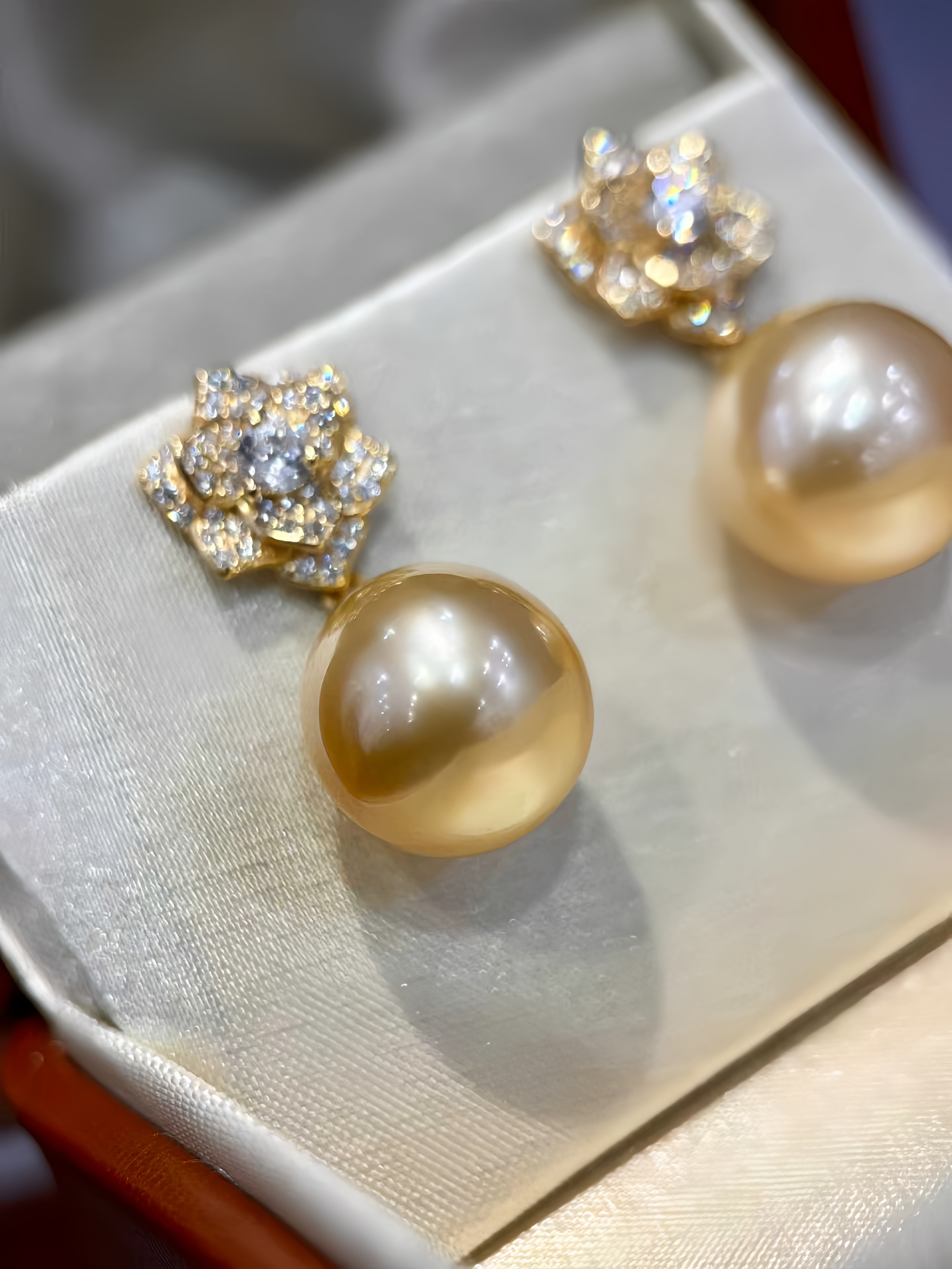 A pair of gold-toned drop earrings with round champagne pearl-like drops and floral diamond settings, displayed on a cream satin jewelry box.