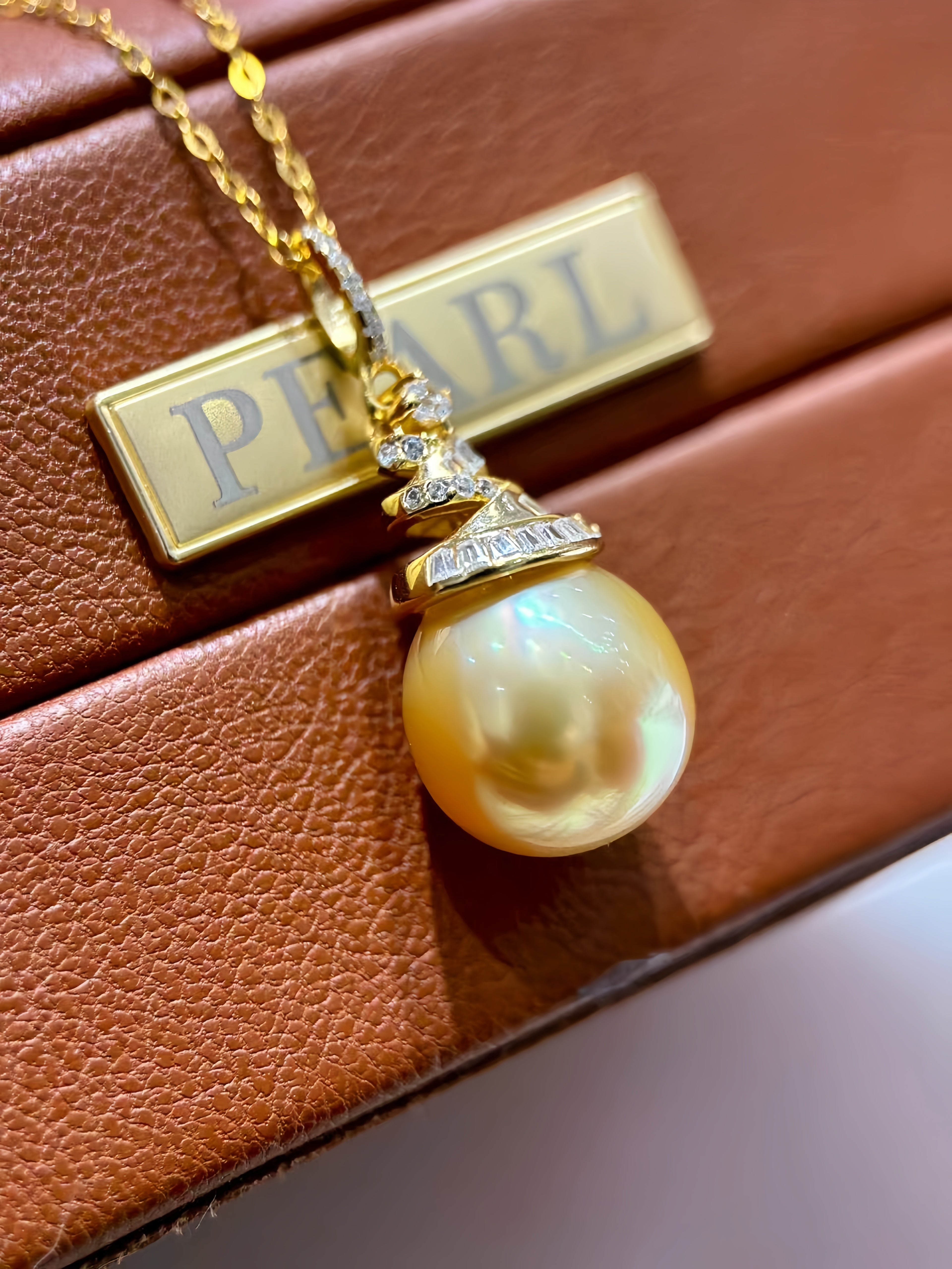 A gold pendant necklace featuring a large, lustrous pearl pendant and a rectangular gold bar engraved with the word 'PEARL', displayed on a brown textured leather surface.