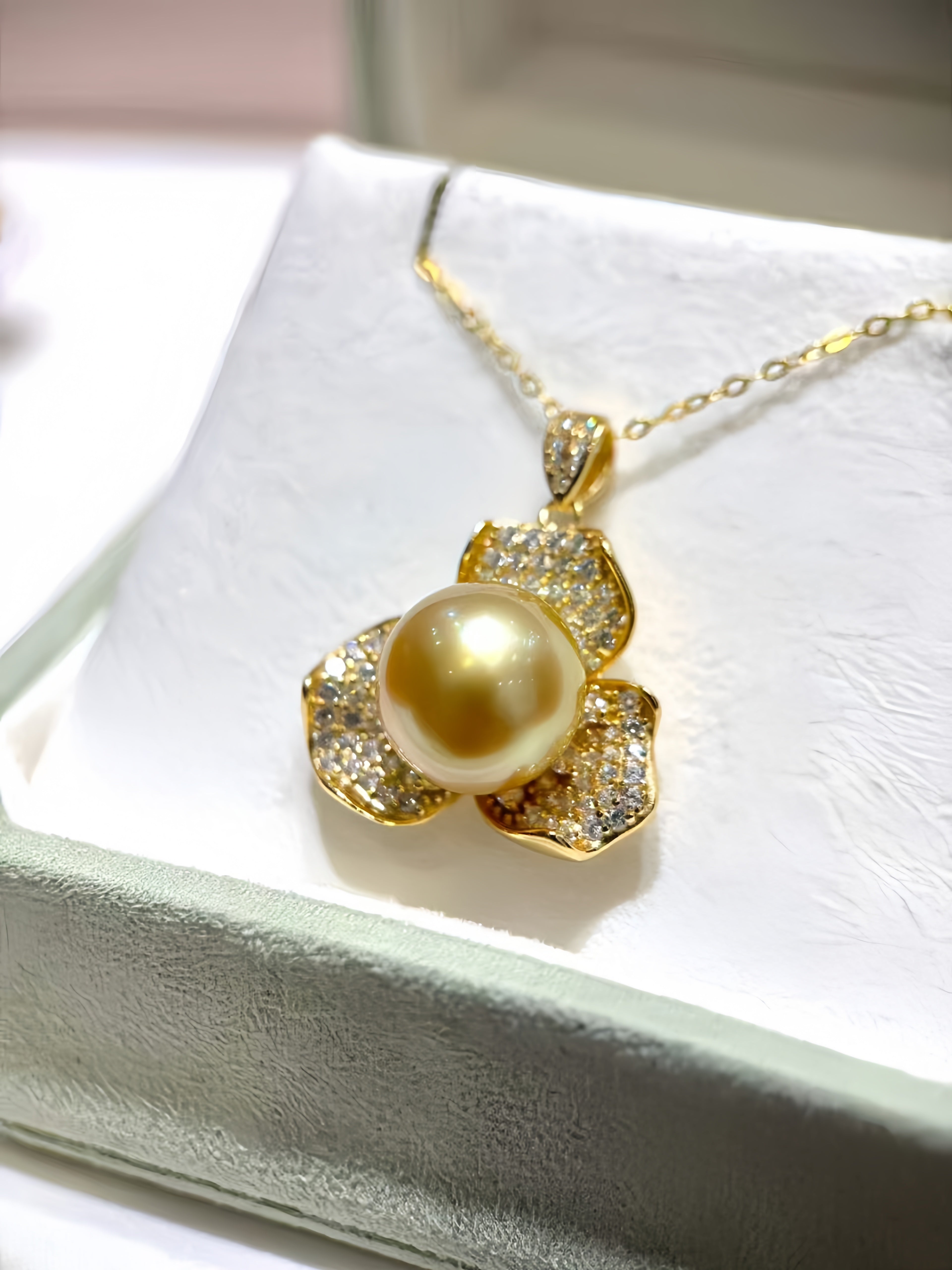 A gold pendant necklace featuring a large round yellow pearl surrounded by five petal-shaped settings paved with small clear stones, displayed on a white jewelry box.