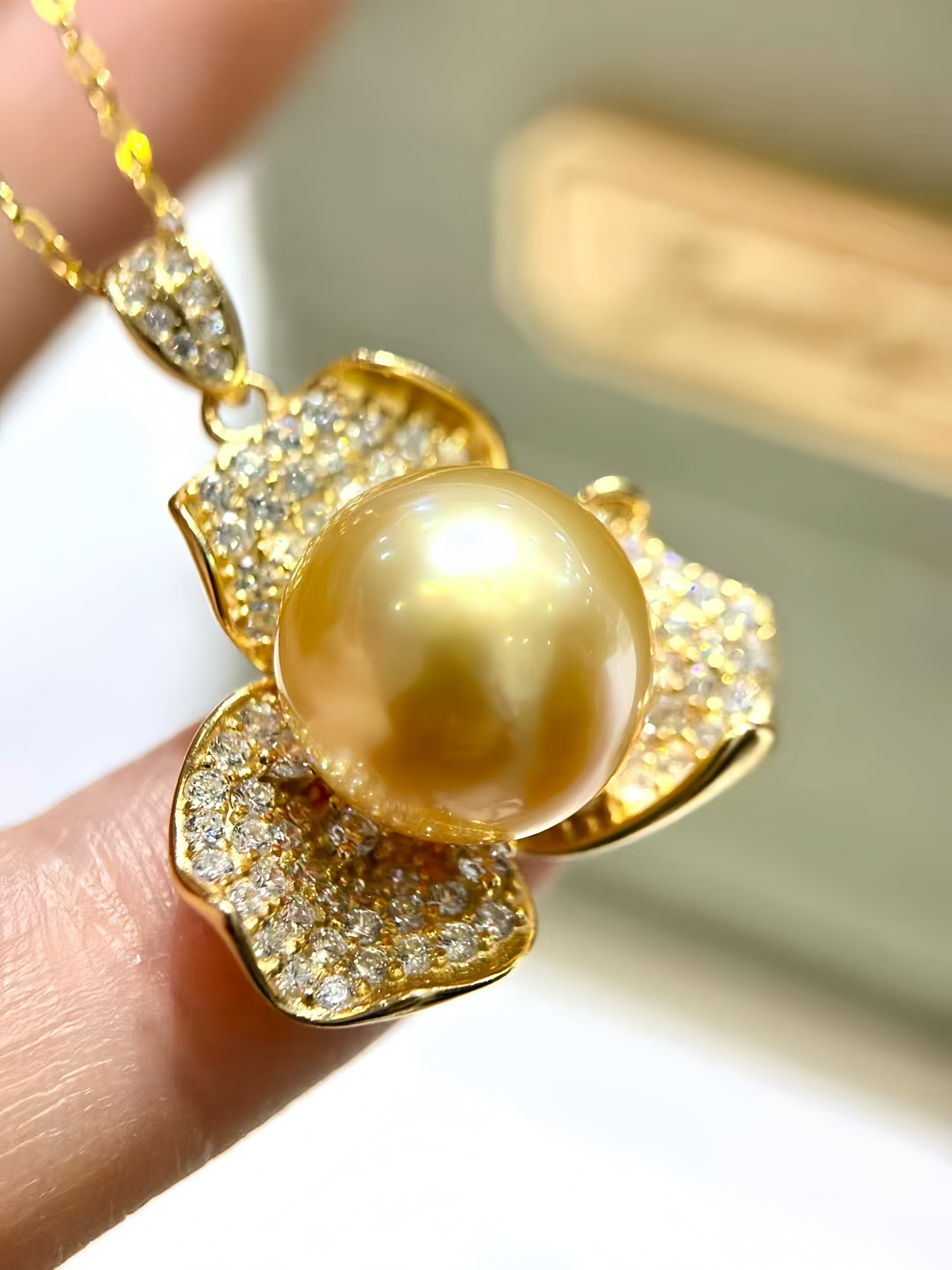 A gold pendant necklace featuring a large round golden pearl surrounded by gold flower-like petals paved with small clear stones, held between fingers against a softly blurred light background.
