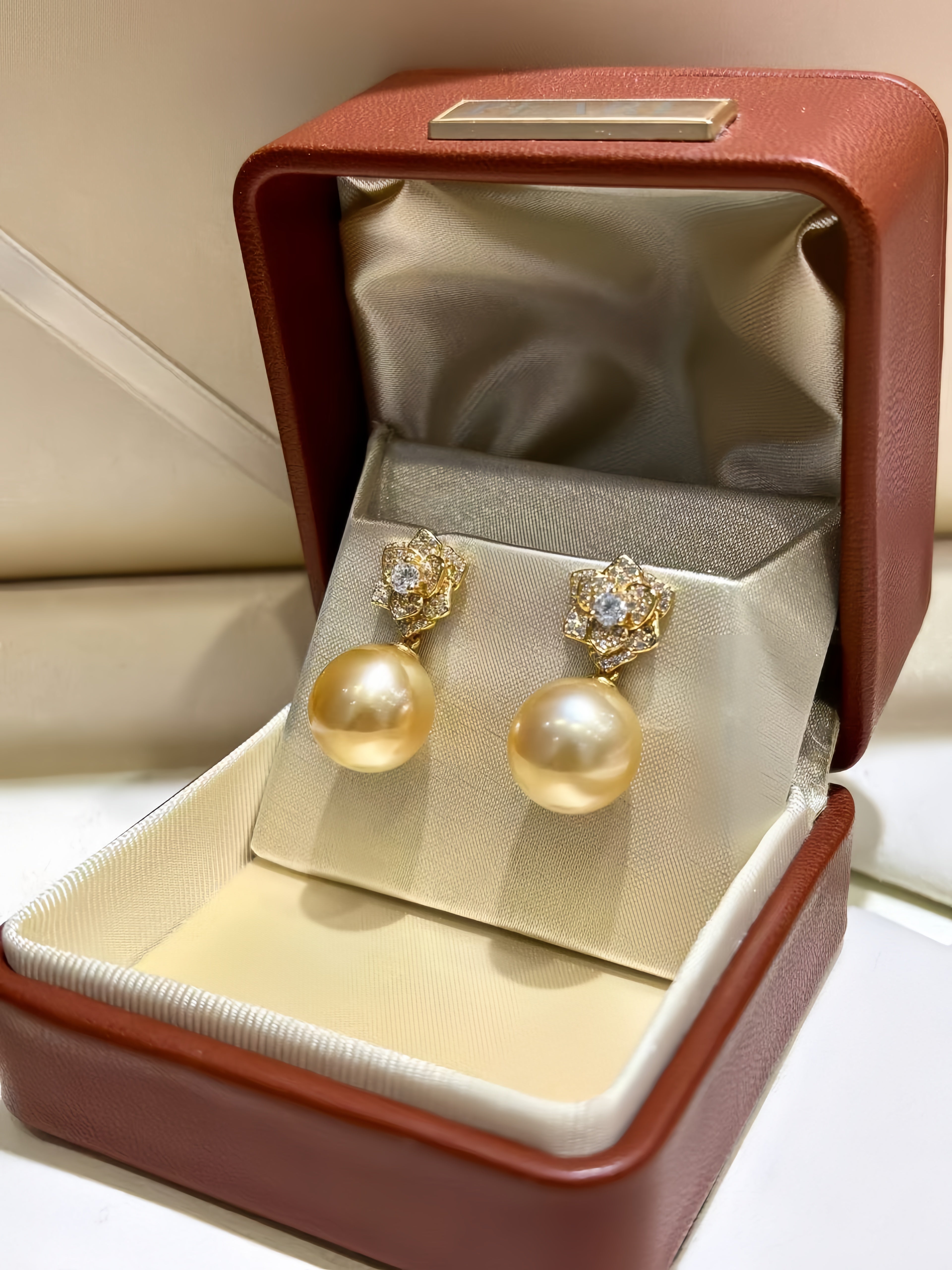 A pair of gold drop earrings with large round pearl-like drops and small clear stones in the tops, displayed on a cream velvet stand inside an open reddish-brown jewelry box.