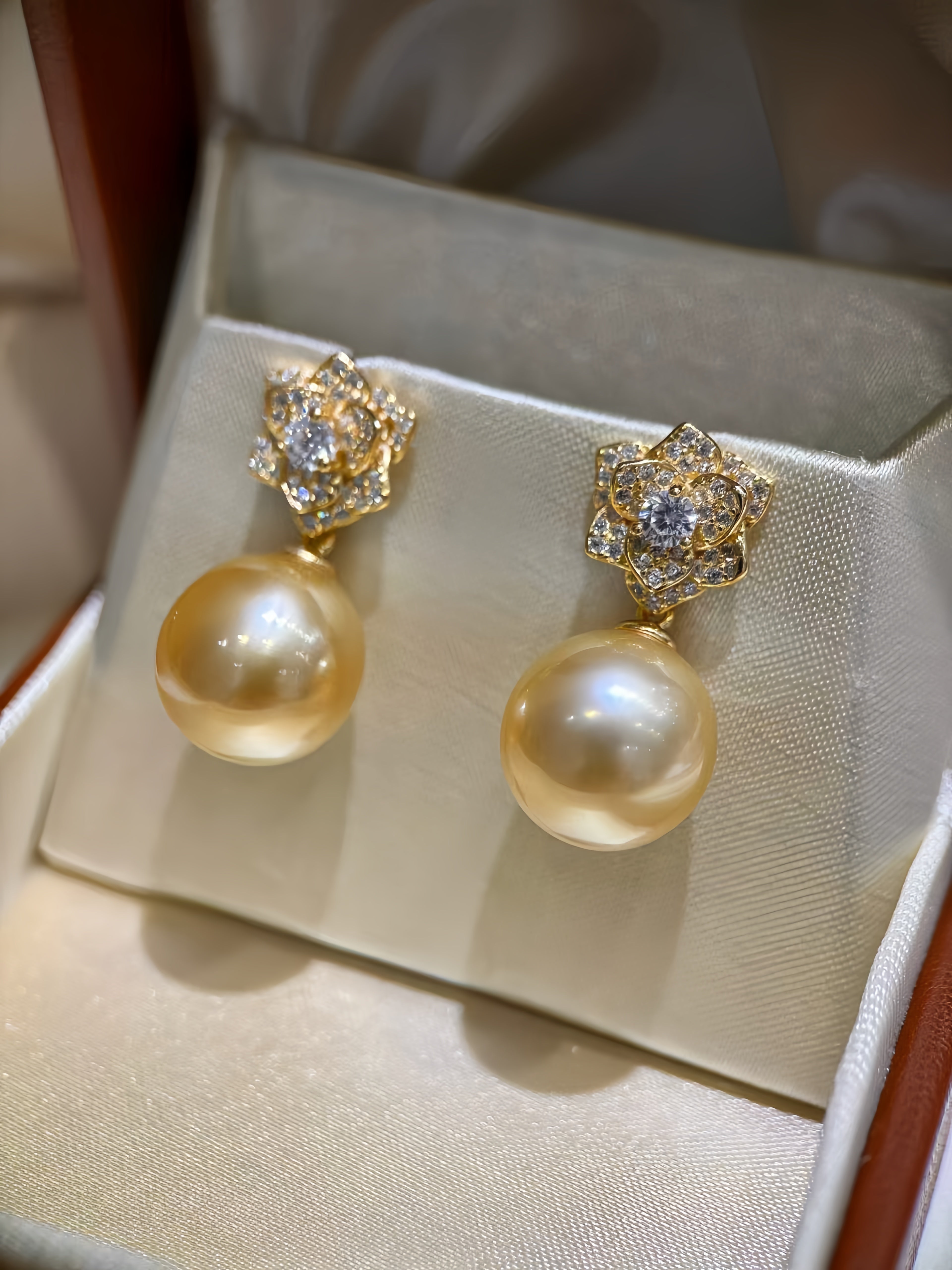 A pair of gold drop earrings with large round cream pearls and floral gold settings with clear stones, displayed on a cream satin jewelry box.