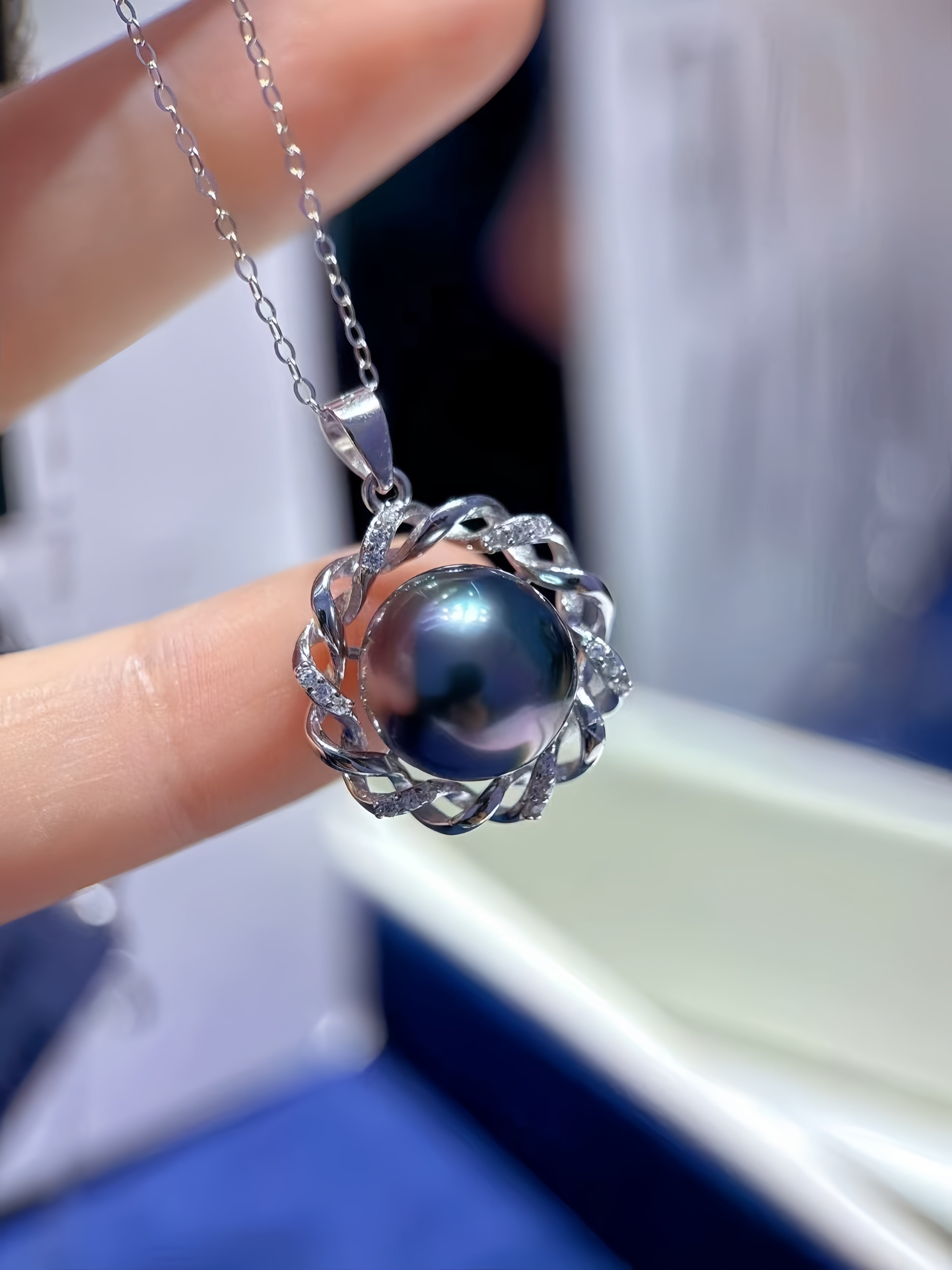 A close-up of a silver pendant necklace with a dark, iridescent pearl held by a hand. The pendant features a silver filigree-like border with small clear stones, set against a blurred background.